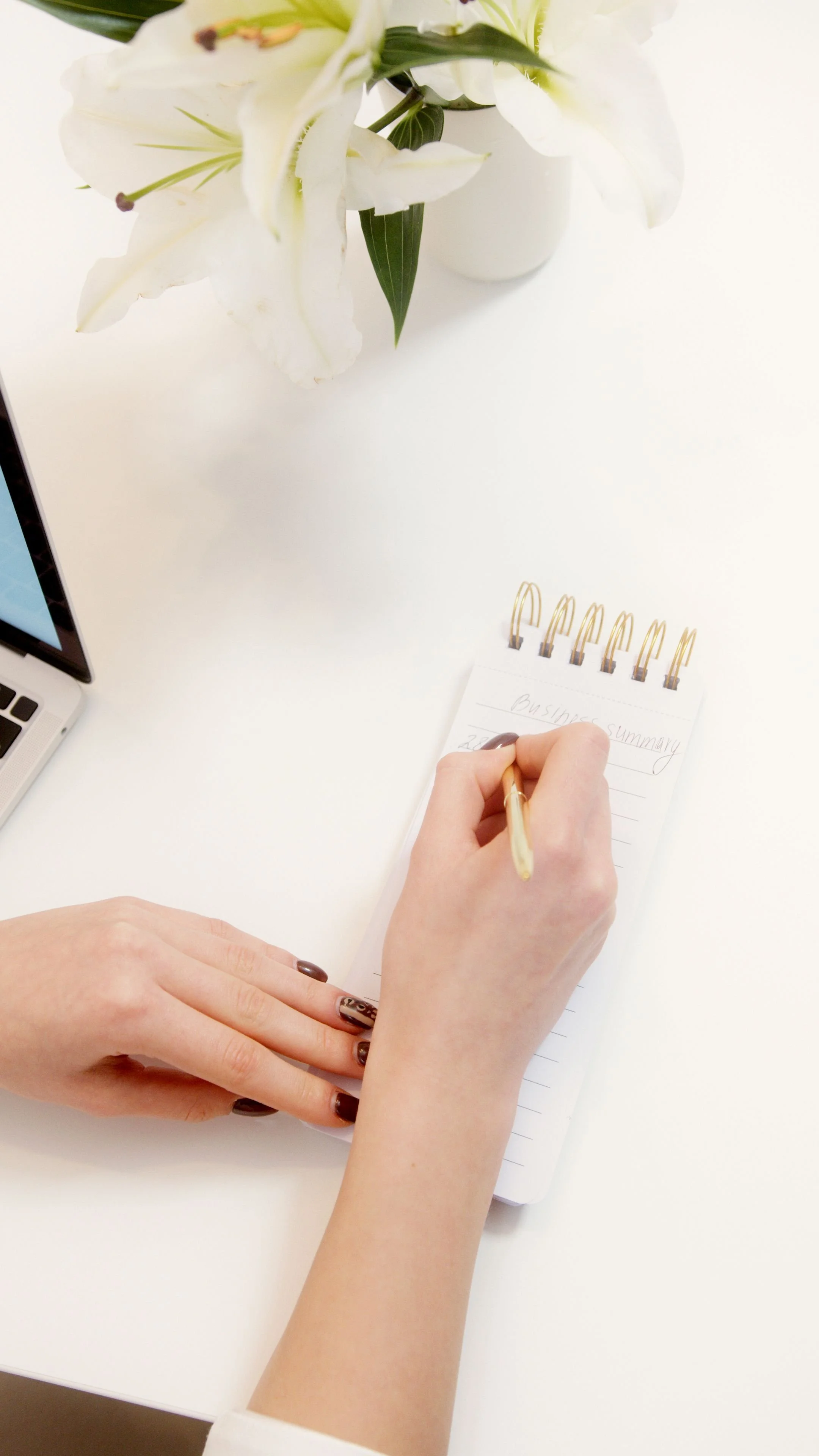 A person's hands writing in a small spiral notebook labeled 'Business Summary' on a white desk. A vase with white flowers and a laptop are also on the desk.