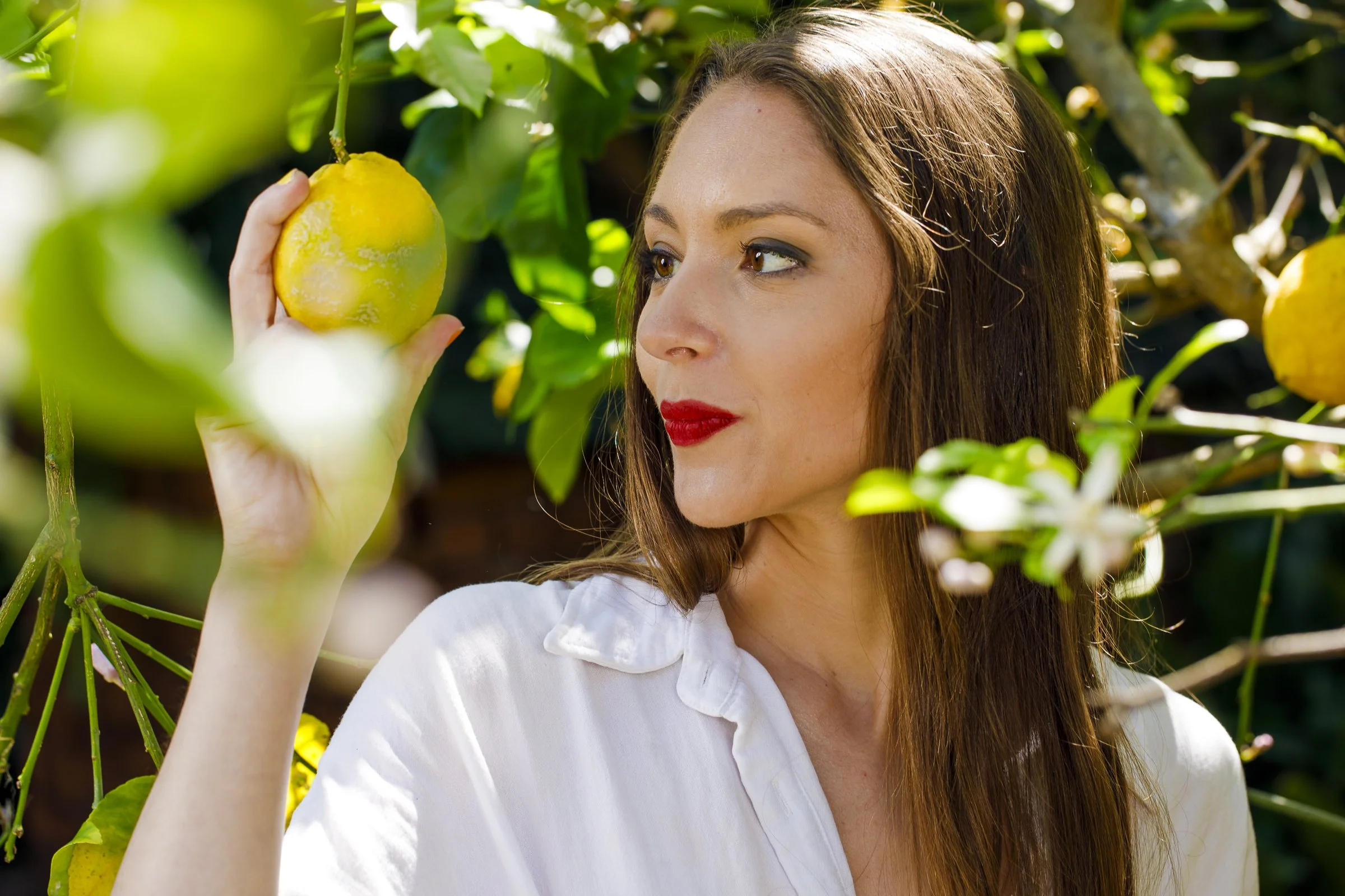 A woman with brown hair and red lipstick holding a yellow lemon on a tree