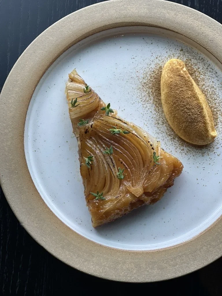 Et stykke av eplekake med tynne skiver av eple, pyntet med friske urter, servert med en pjusk av kanel eller krydder på en hvit tallerken med en beige kant.