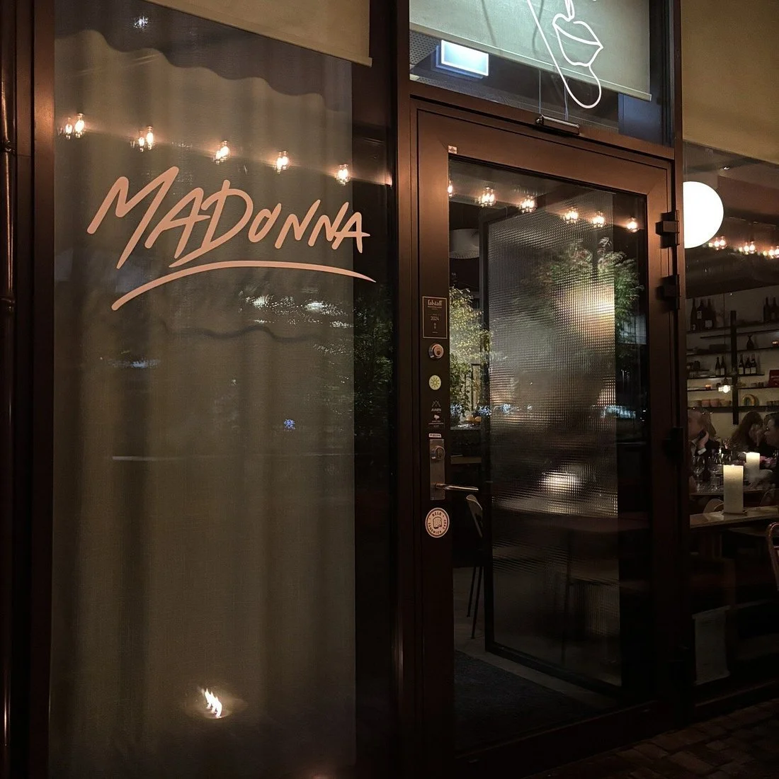 Utsiden av en restaurant kalt Madonna med glassdør og vindu, dekorert med neonlys og lamper inne, med et stearinlys på bordet inne og noen mennesker som spiser.