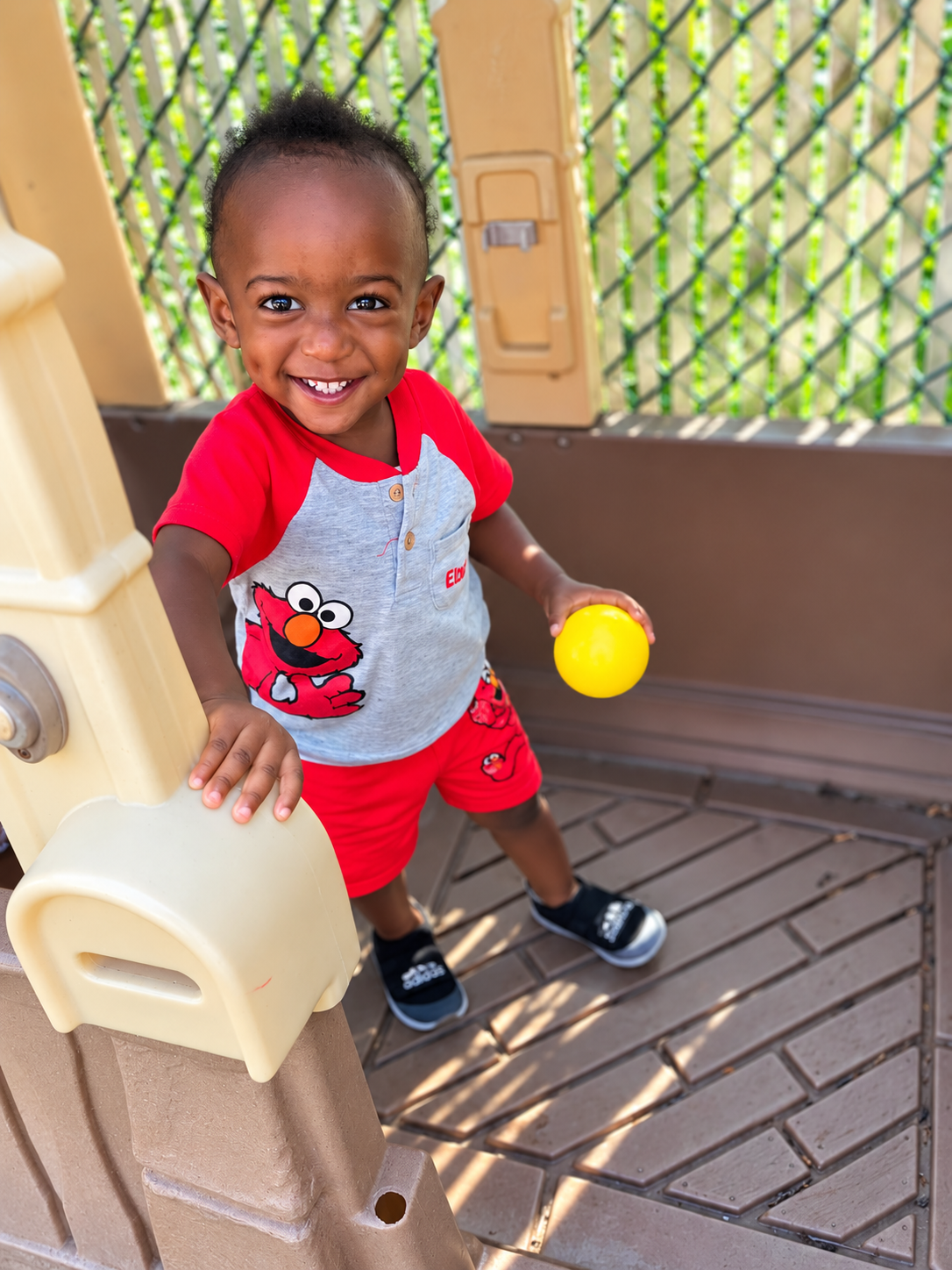 Smiling young boy holding a yellow ball, wearing a gray and red Elmo shirt and shorts, standing on a wooden deck at a playground.
