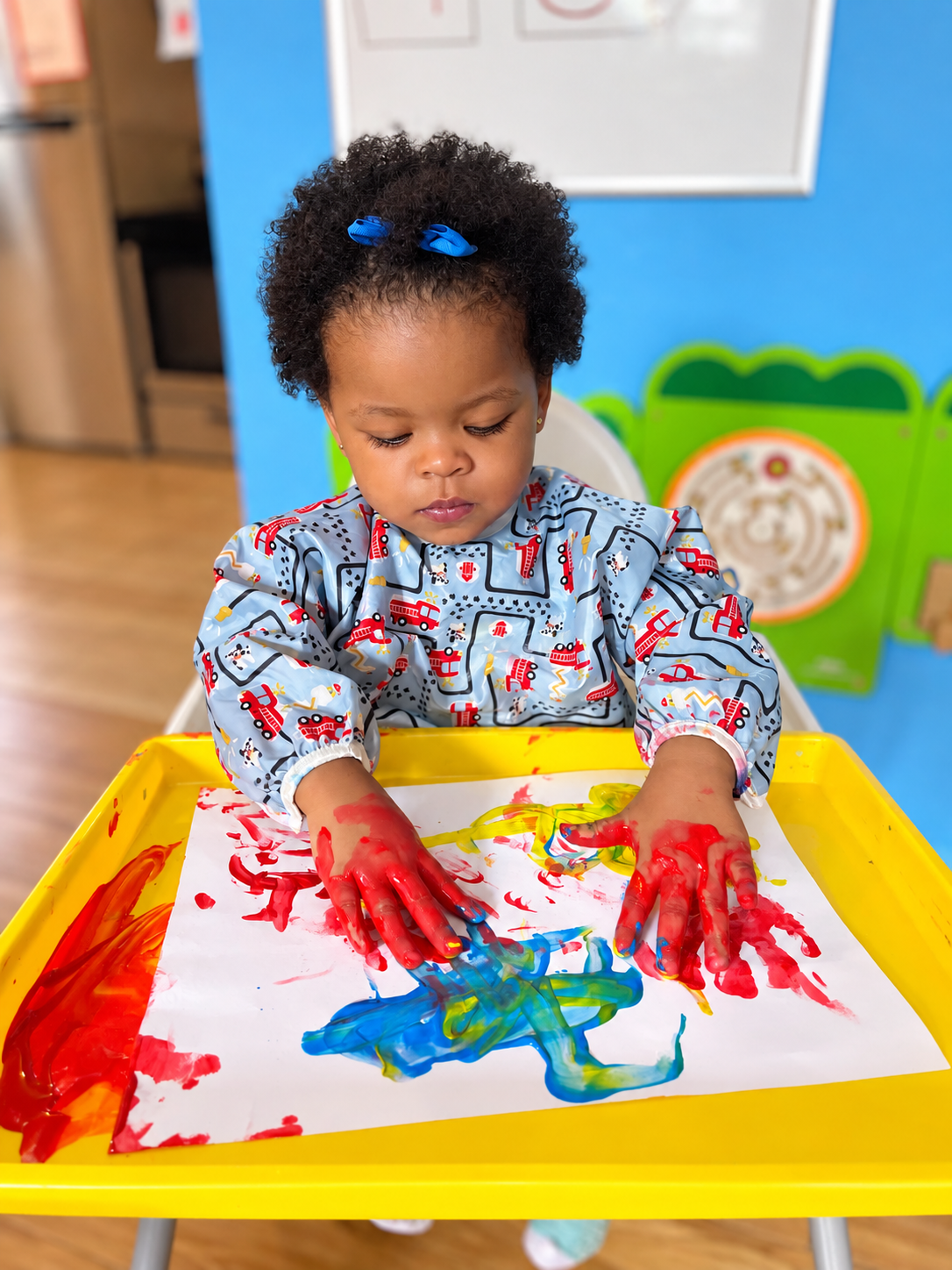 A young girl with curly hair and a blue bow is engaging in finger painting with red, yellow, and blue paint on a sheet of paper in a yellow tray, in a colorful indoor setting.