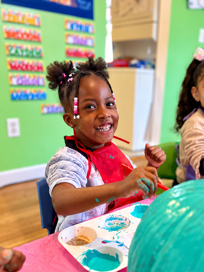 A young girl with braided hair and beads, smiling while painting at a table, wearing a red apron in a colorful classroom.