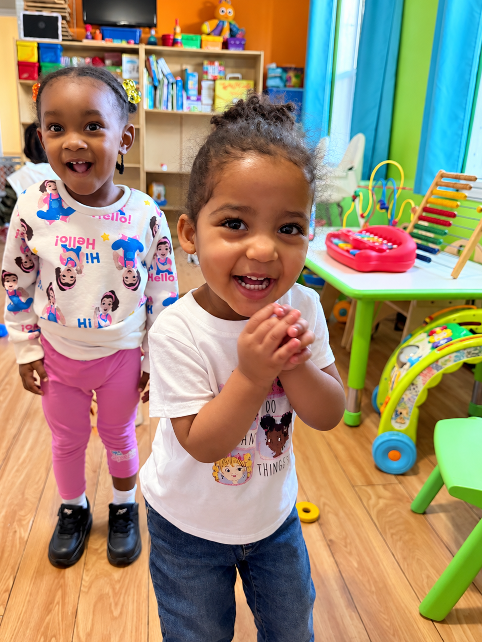 Two young girls smiling happily in a colorful classroom with toys and books in the background.