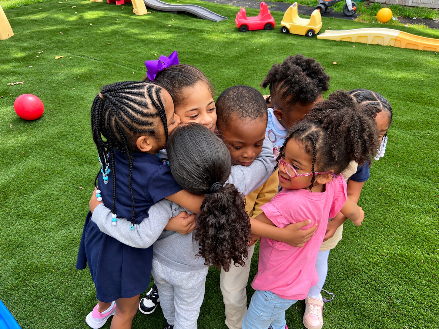 Group of seven children of diverse ethnicities sharing a group hug on a green outdoor playground with toys and a slide in the background.