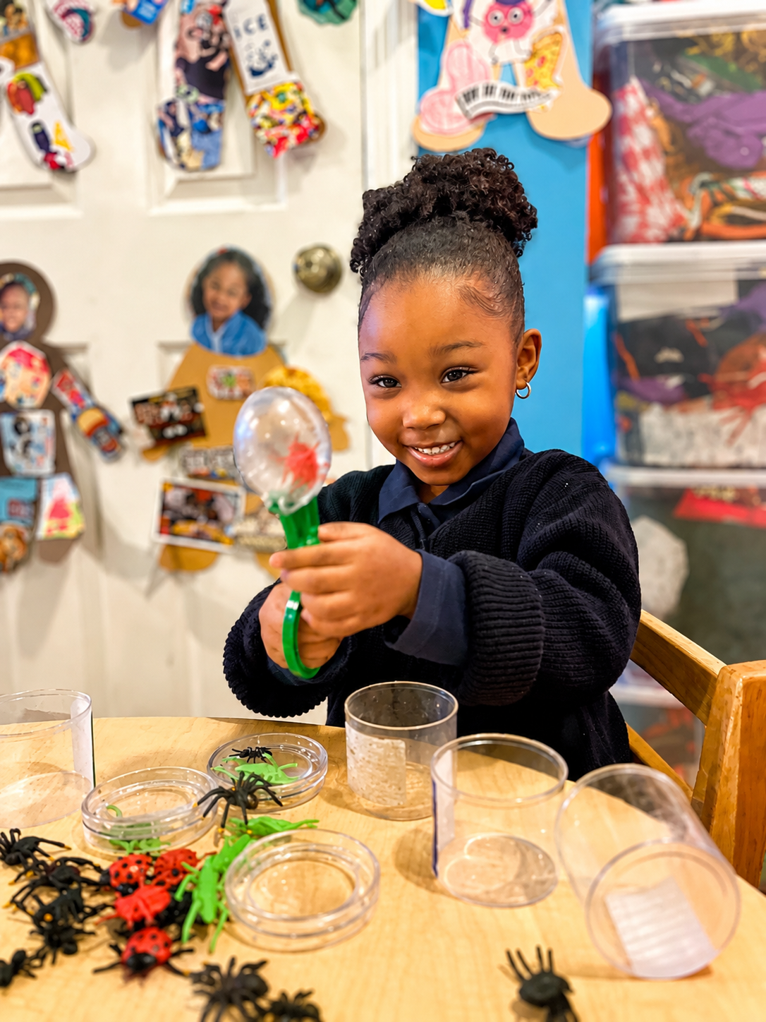 A smiling young girl with curly hair in a bun holds a small toy bubble wand. She is sitting at a wooden table with various toy insects, including spiders, ladybugs, and centipedes. The background has colorful decorations and pictures on a white door.