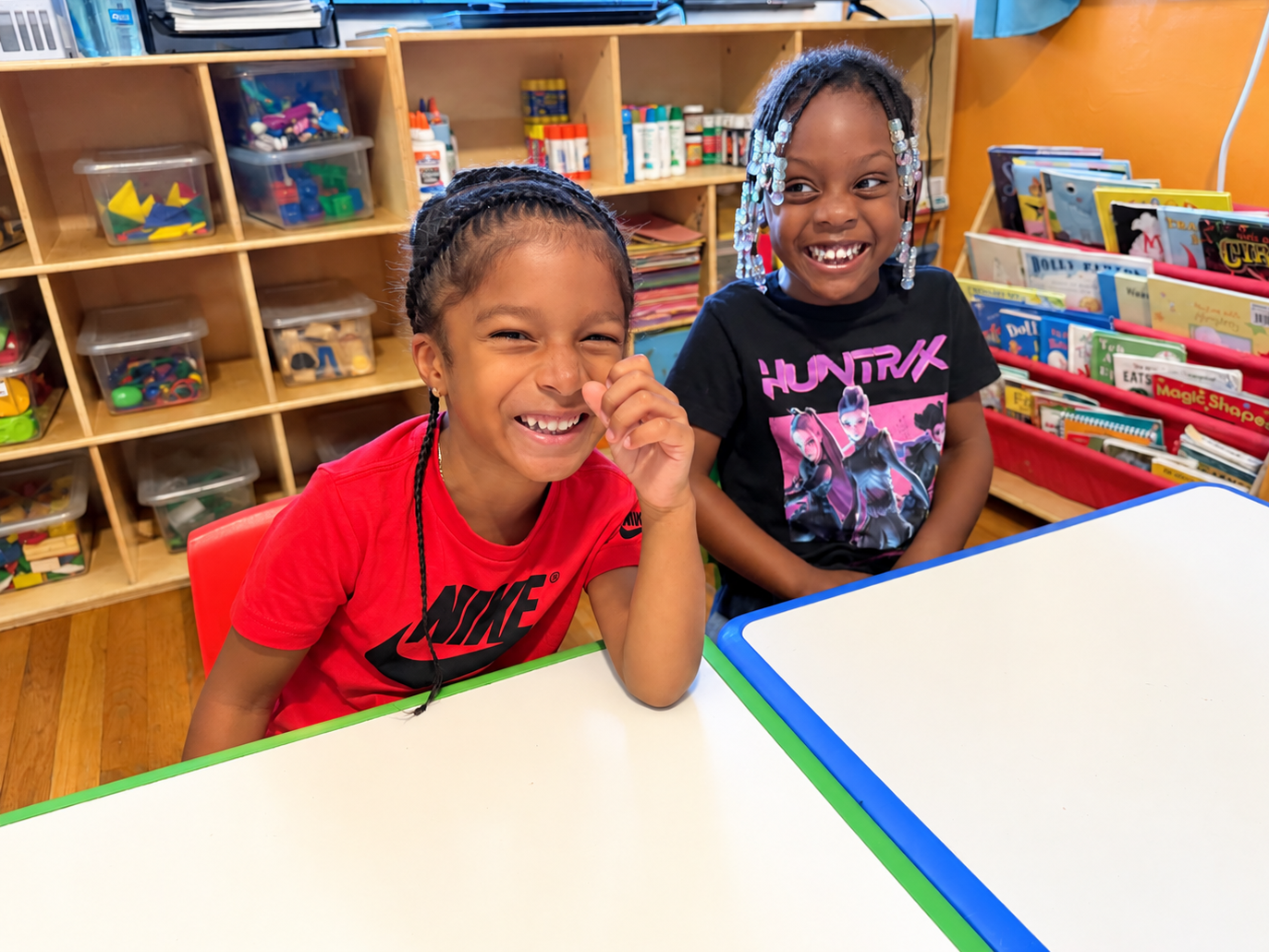 Two young kids smiling and sitting at a classroom table, with shelves filled with books, art supplies, and toys in the background.