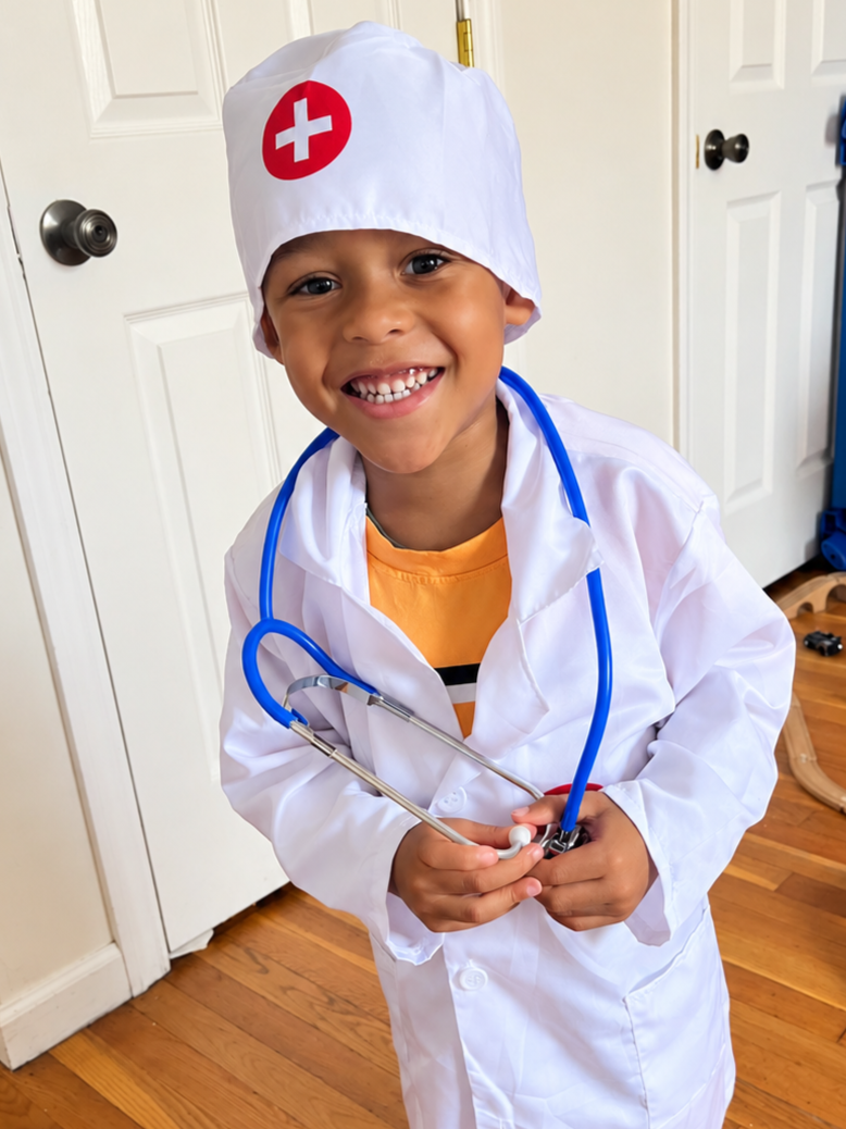 A young boy dressed as a doctor wearing a white coat, a nurse hat with a red cross, and holding a toy stethoscope, smiling in a room with wooden floor and white doors.