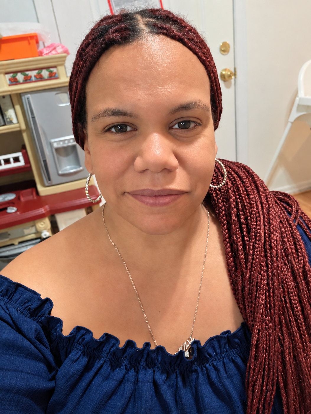 A woman with braided red hair wearing a blue off-shoulder top, silver hoop earrings, and a silver necklace with a pendant, smiling at the camera in a room with kitchen shelves and a toy kitchen set in the background.
