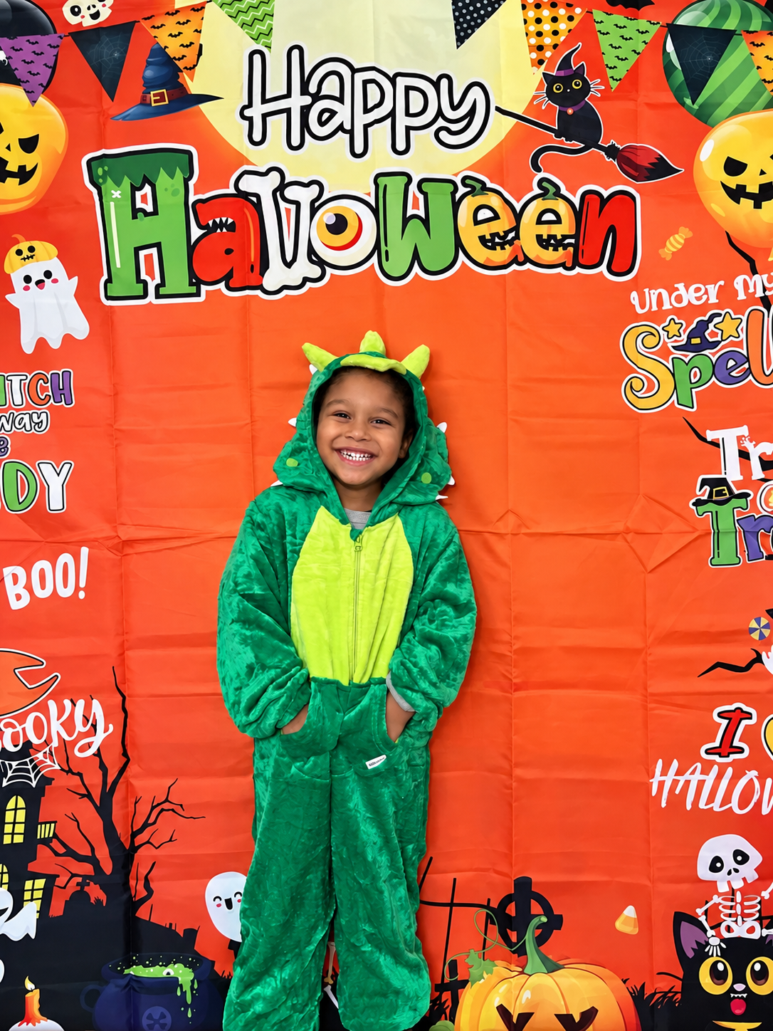 A young boy dressed in a green dinosaur costume with a hood featuring dinosaur eyes and horns, smiling against a festive Halloween backdrop with pumpkins, ghosts, witches, and colorful lettering.