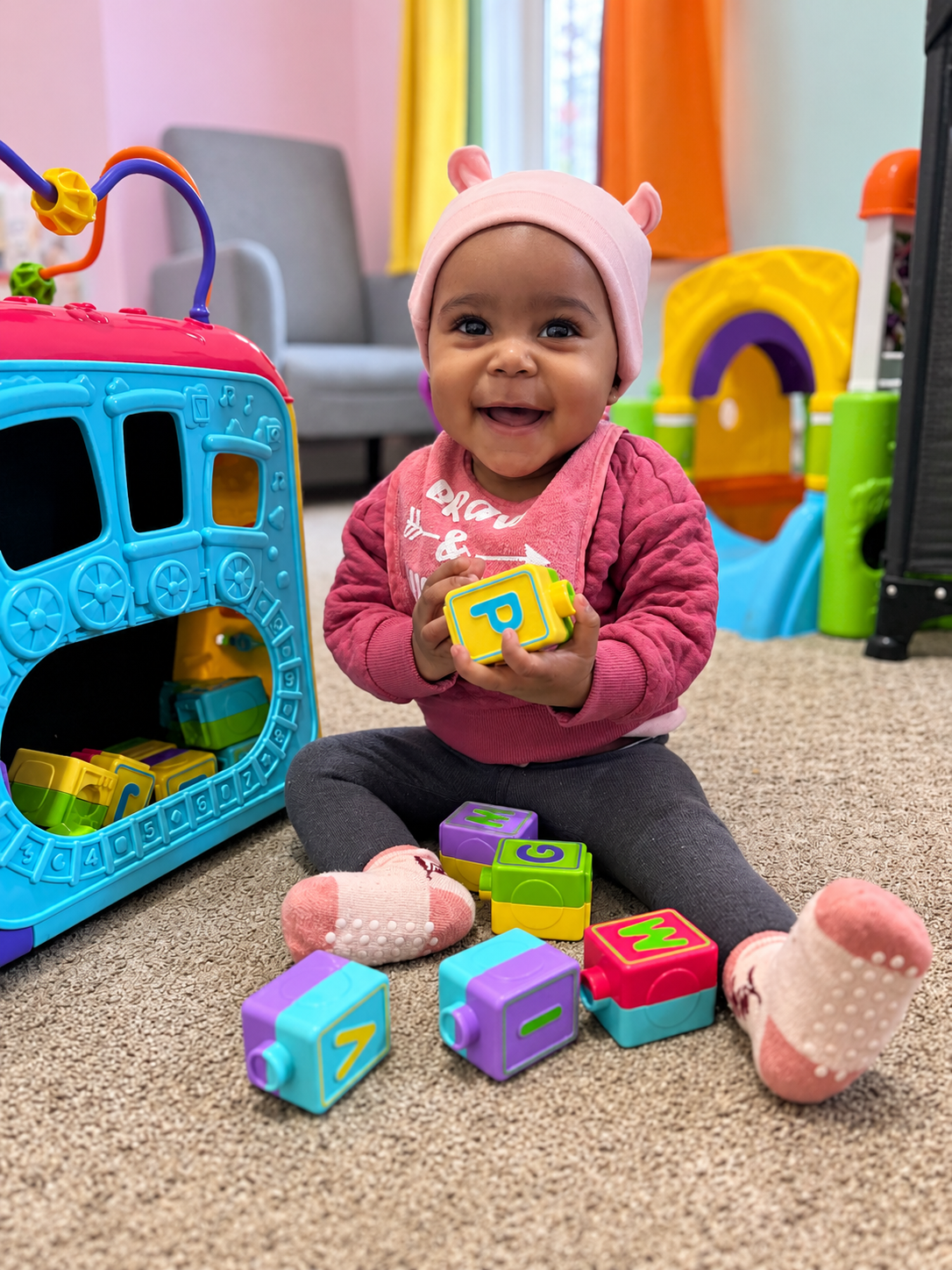 Smiling infant girl wearing a pink hat and pink sweatshirt, sitting on carpeted floor surrounded by colorful alphabet blocks and plush toys, playing in a room with vibrant curtains and toy structures.