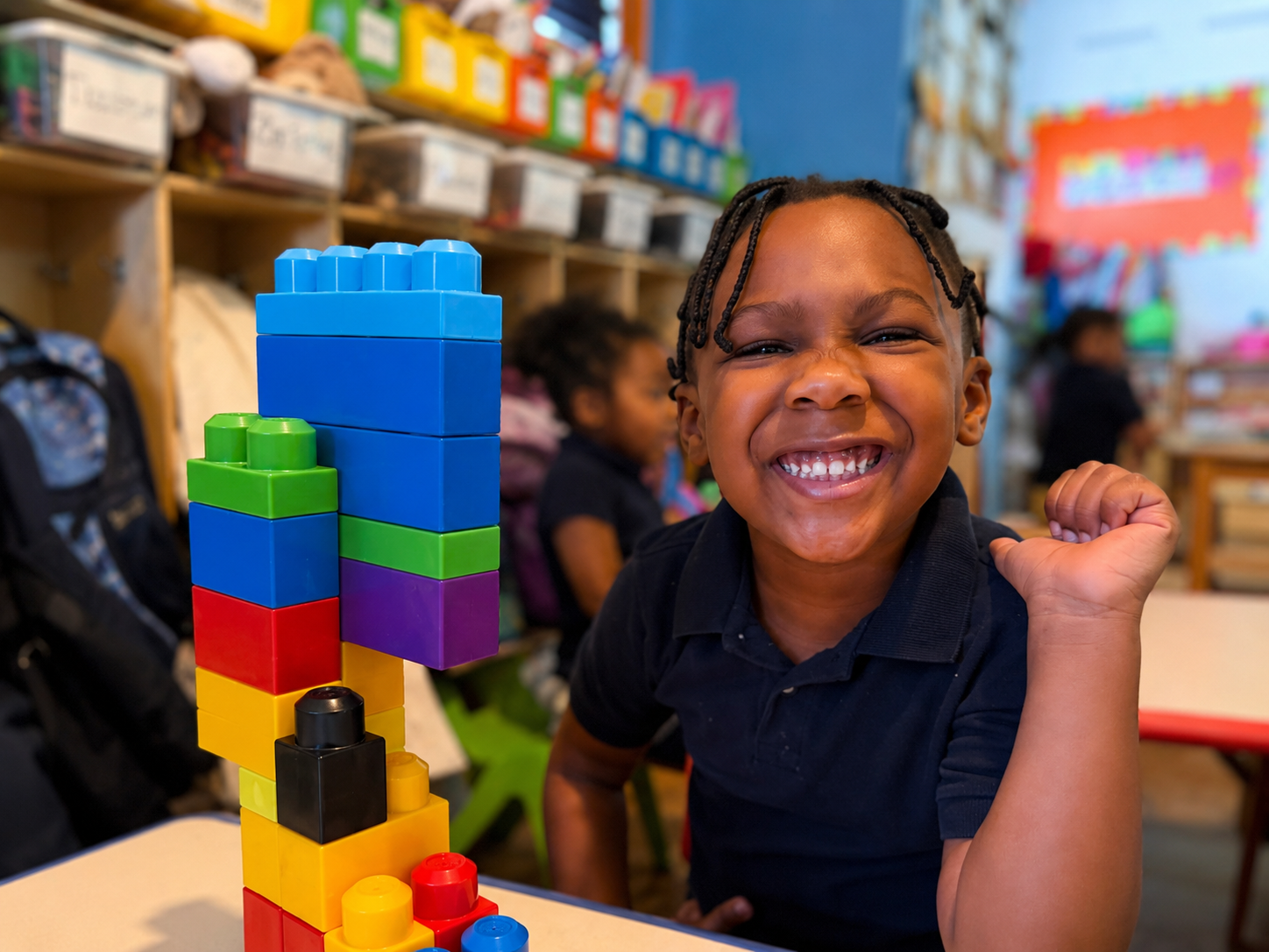A young boy in a classroom smiling proudly playing with colorful stack of building blocks.