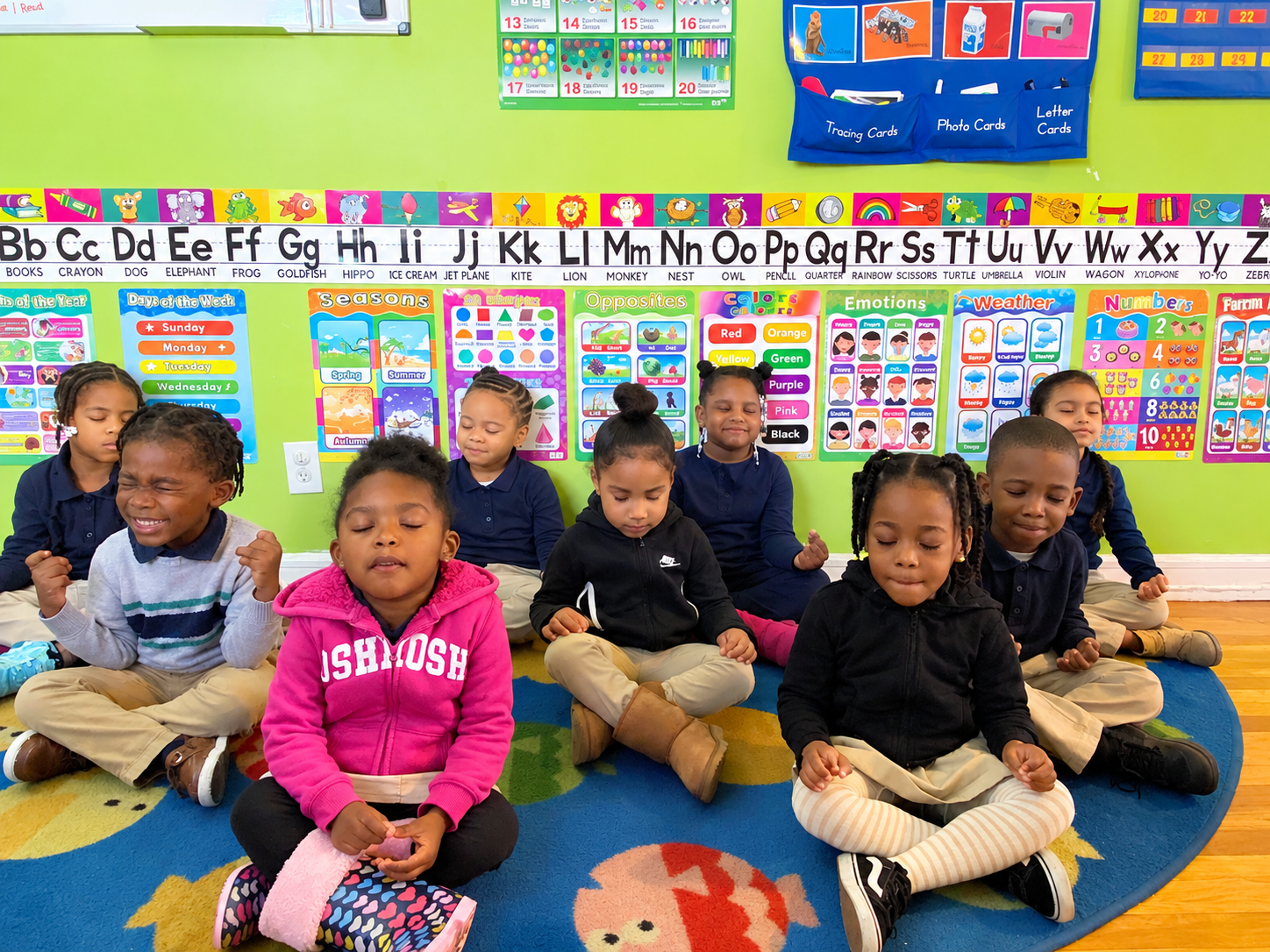 A group of young children sitting cross-legged on a colorful classroom rug with their eyes closed, appearing to meditate or relax. The classroom has brightly colored educational posters and charts on the walls behind them.