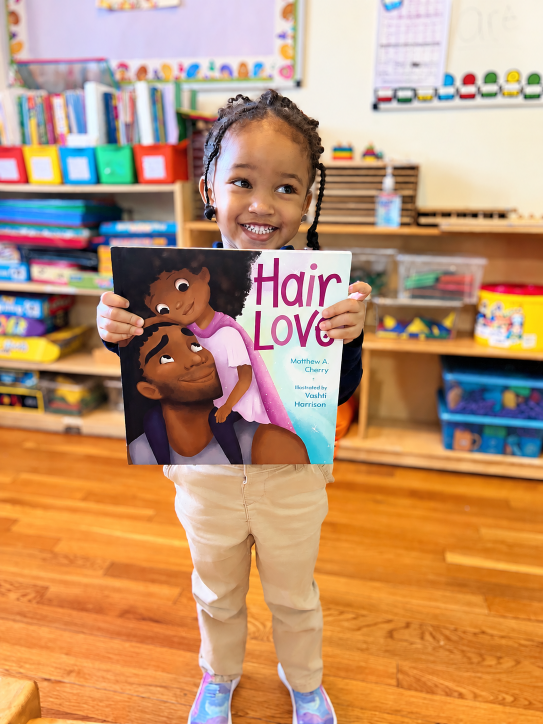 A young girl smiling in a classroom holding a children's book titled 'Hair Love' with an illustration of two children with curly hair on the cover.