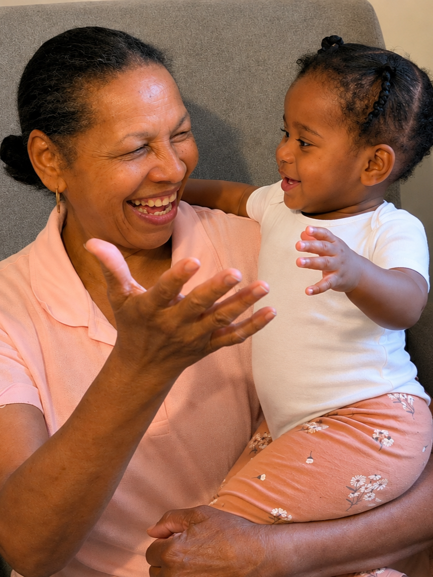 An elderly woman and a young girl laughing and playing together, sitting on a gray chair, sharing a joyful moment.