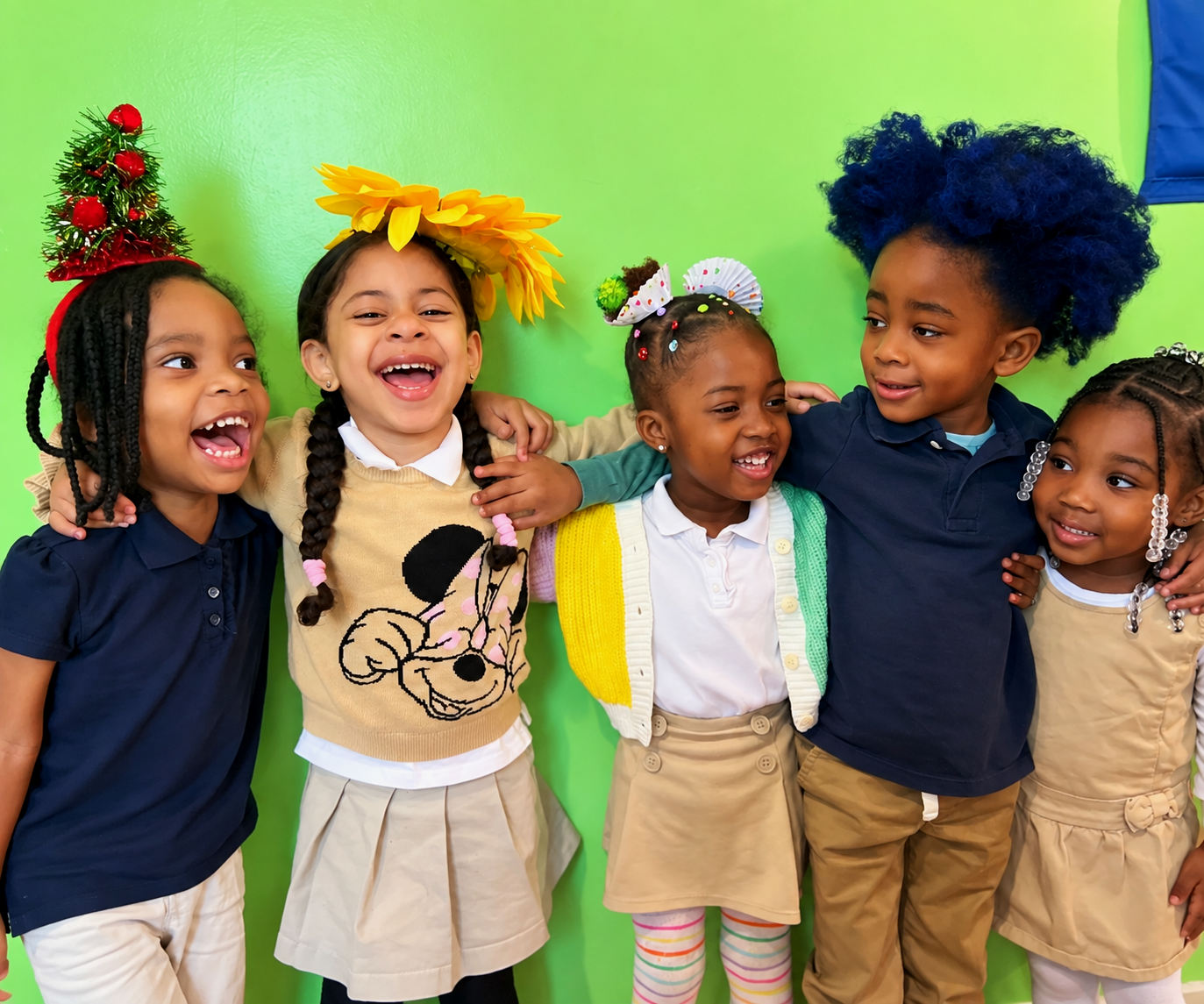 Group of six diverse children smiling, standing with arms around each other against a green wall, celebrating. Some wear festive accessories like a Christmas tree hat, sunflower headband, and colorful hair decorations.