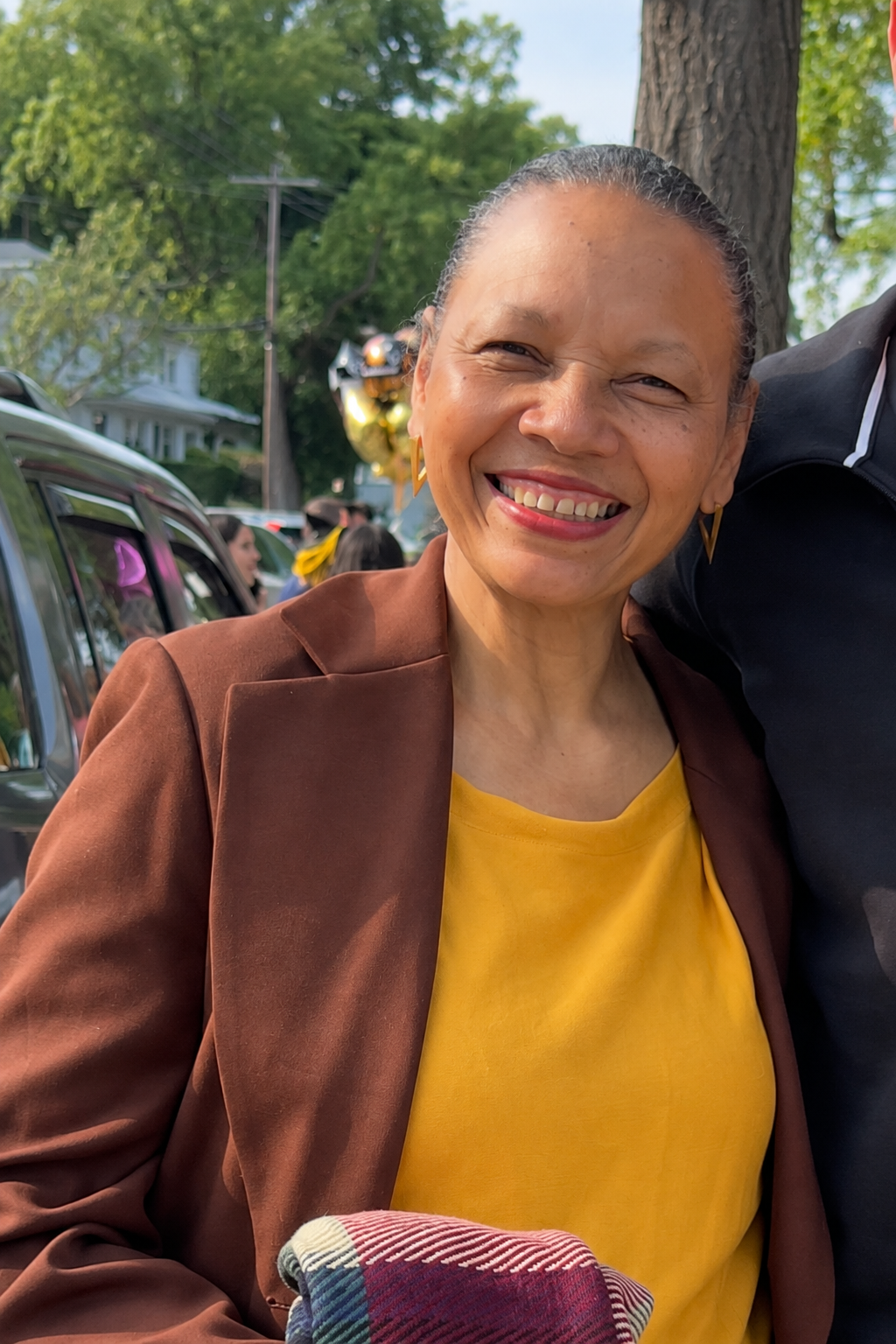 Smiling woman with dark hair tied back, wearing a yellow top and brown blazer, standing outside with trees, balloons, and several people in the background.