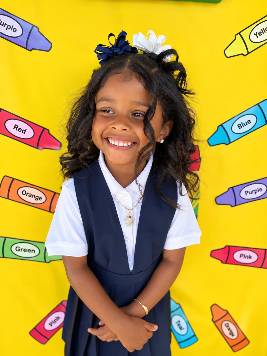 A young girl with curly black hair, adorned with blue and white bows, smiling in front of a colorful background with crayon drawings labeled with colors.