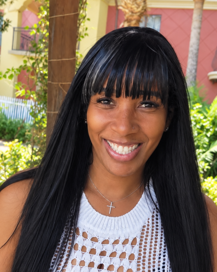 Smiling woman with long black hair, wearing a white crochet top, a cross necklace, and small earrings, outdoors with trees and a colorful building in the background.