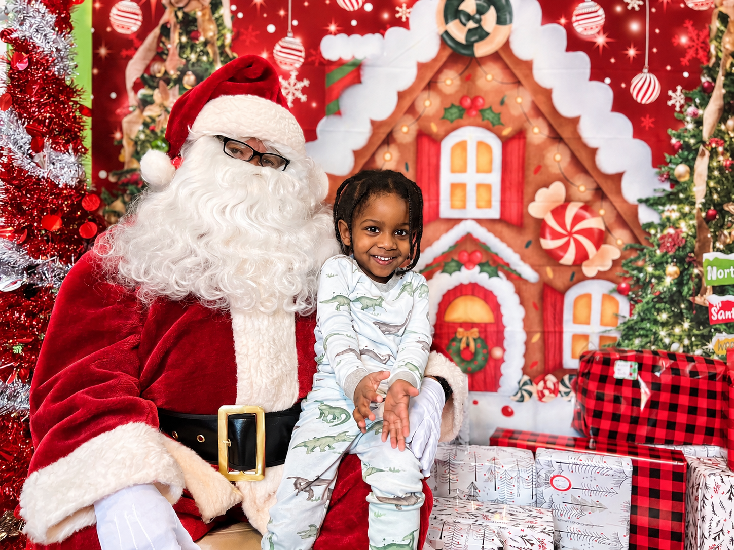 A young boy sitting with Santa during a Christmas event, with holiday-themed decorations and wrapped presents in the background.