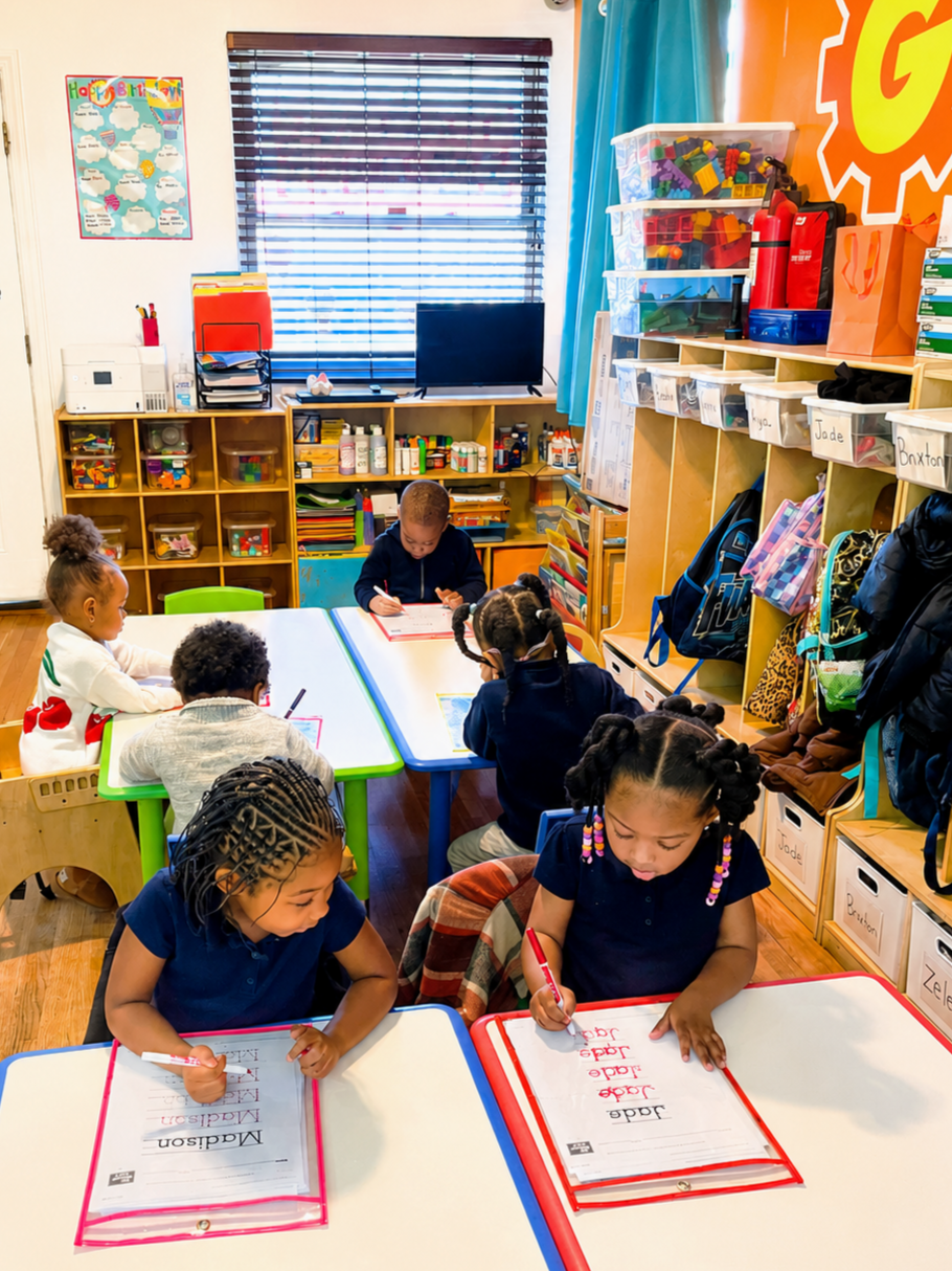 Young children in a classroom sitting at tables, writing on worksheets, with cubbies, colorful storage bins, and educational posters in the background.