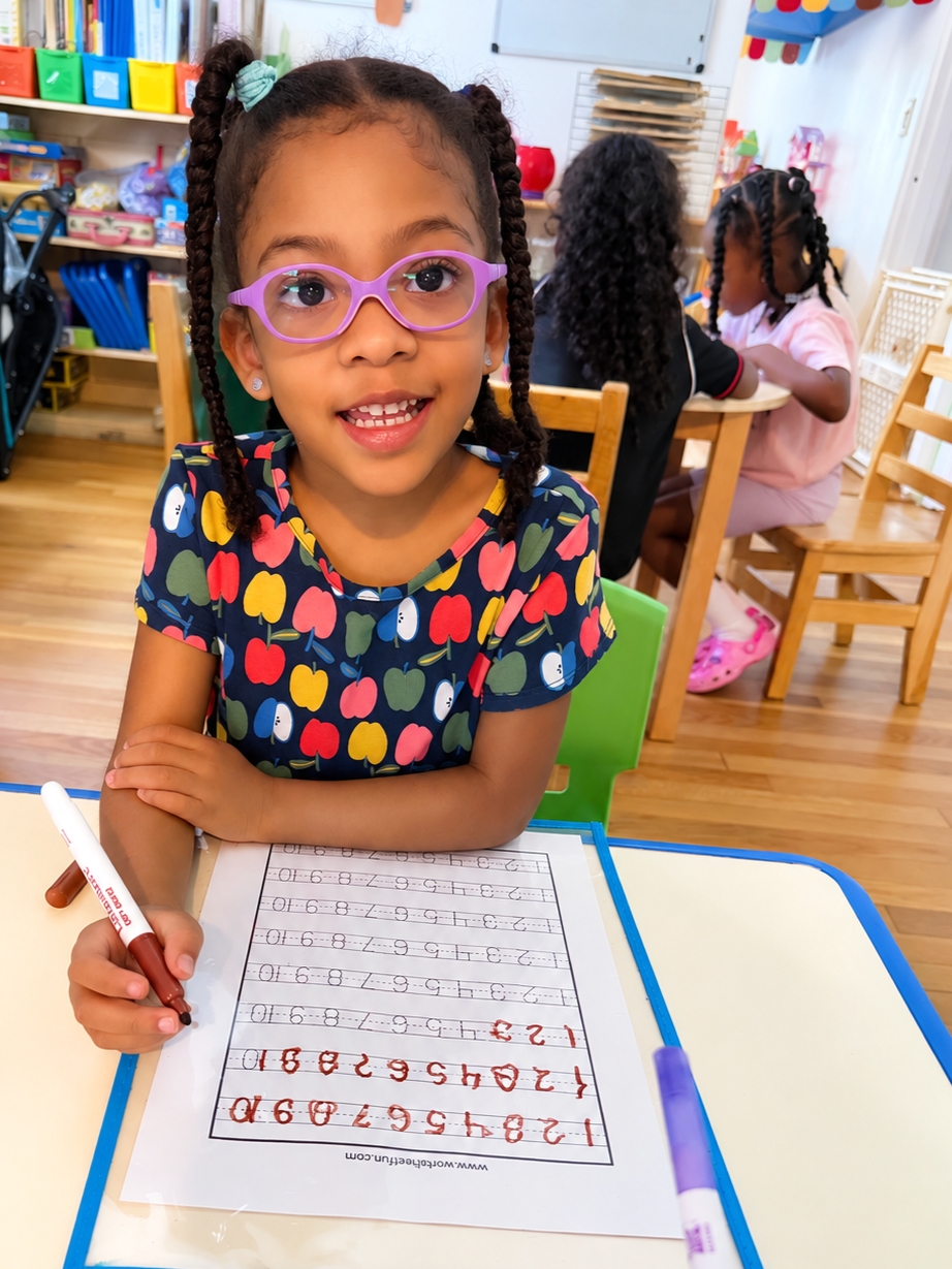 A young girl with glasses and braided hair smiling at the camera while doing handwritten number practice on a sheet of paper in a classroom.