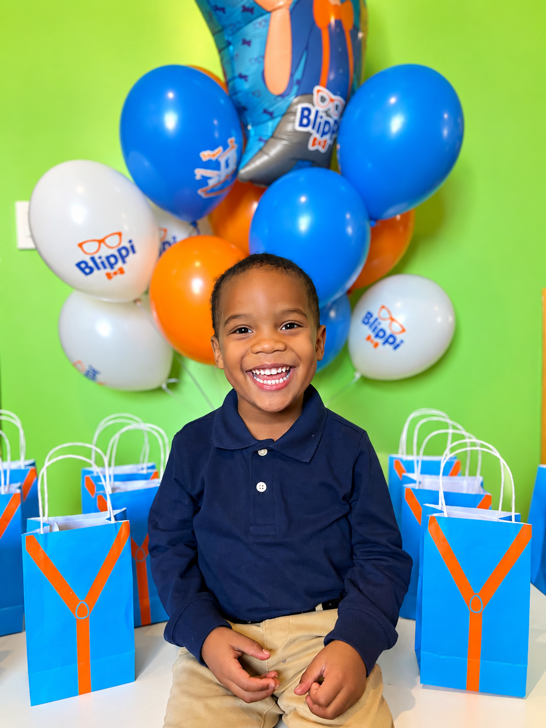 A young boy sitting in front of a birthday celebration backdrop with balloons and gift bags, smiling happily.