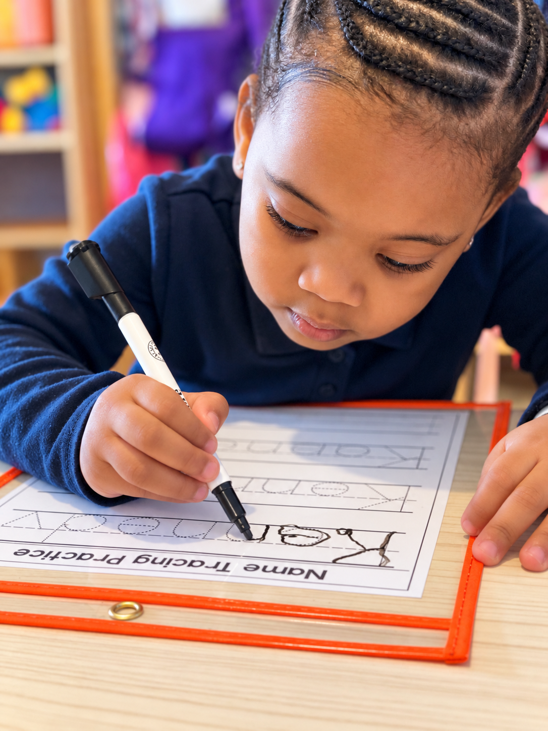 A young girl with braided hair practicing handwriting at a desk, writing the word 'LOVE' with a marker on a paper titled 'Name Tracing Practice'.
