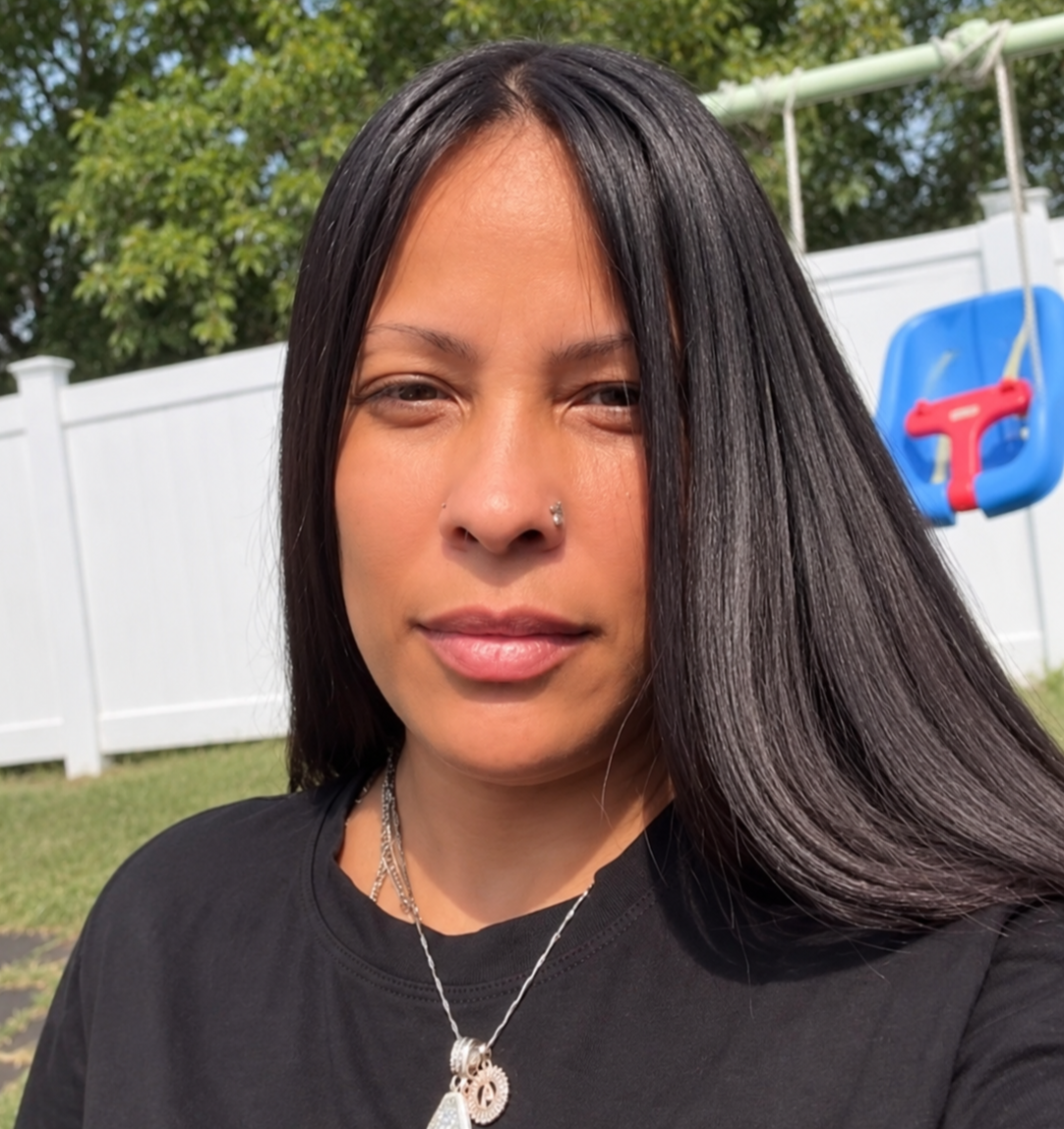 A woman with long black hair, wearing a black shirt and silver jewelry, smiling in a backyard with a white fence and a blue and red swing set in the background.