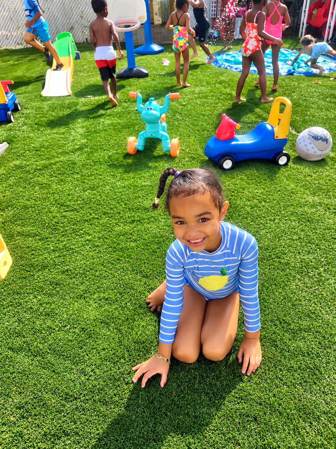 Child smiling and kneeling on grass at a playground, with other children playing and toys scattered around, including a slide, a blue toy car, and a ball.