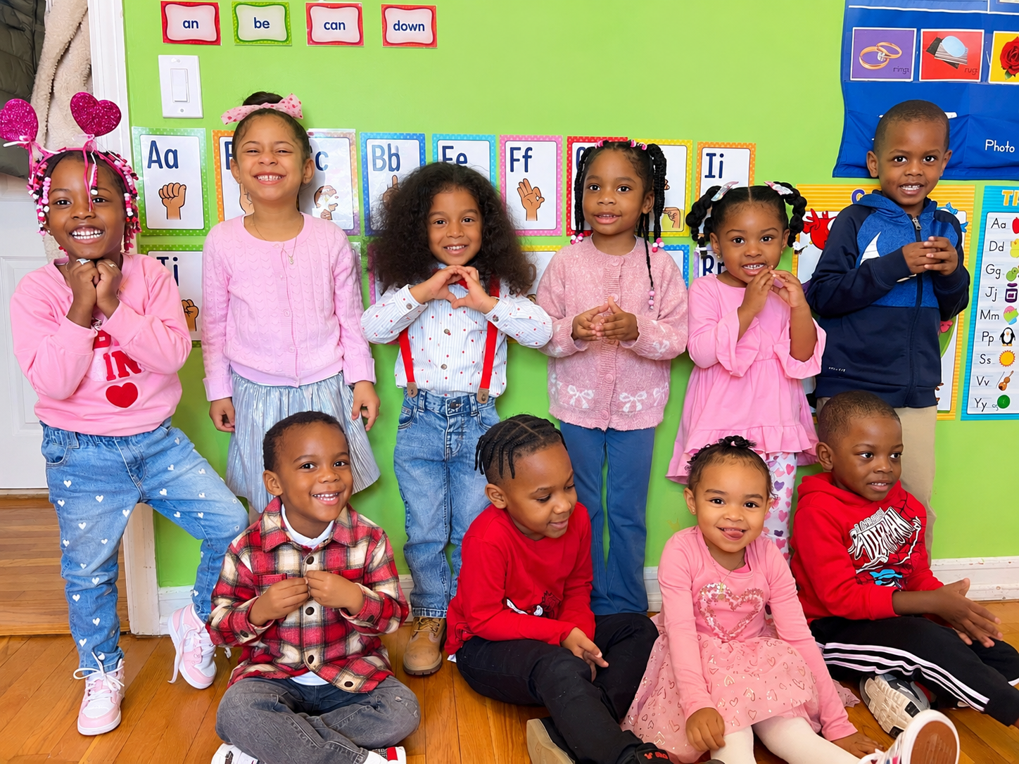 Group of ten diverse young children posing and smiling in a classroom with colorful educational posters and wall decorations.