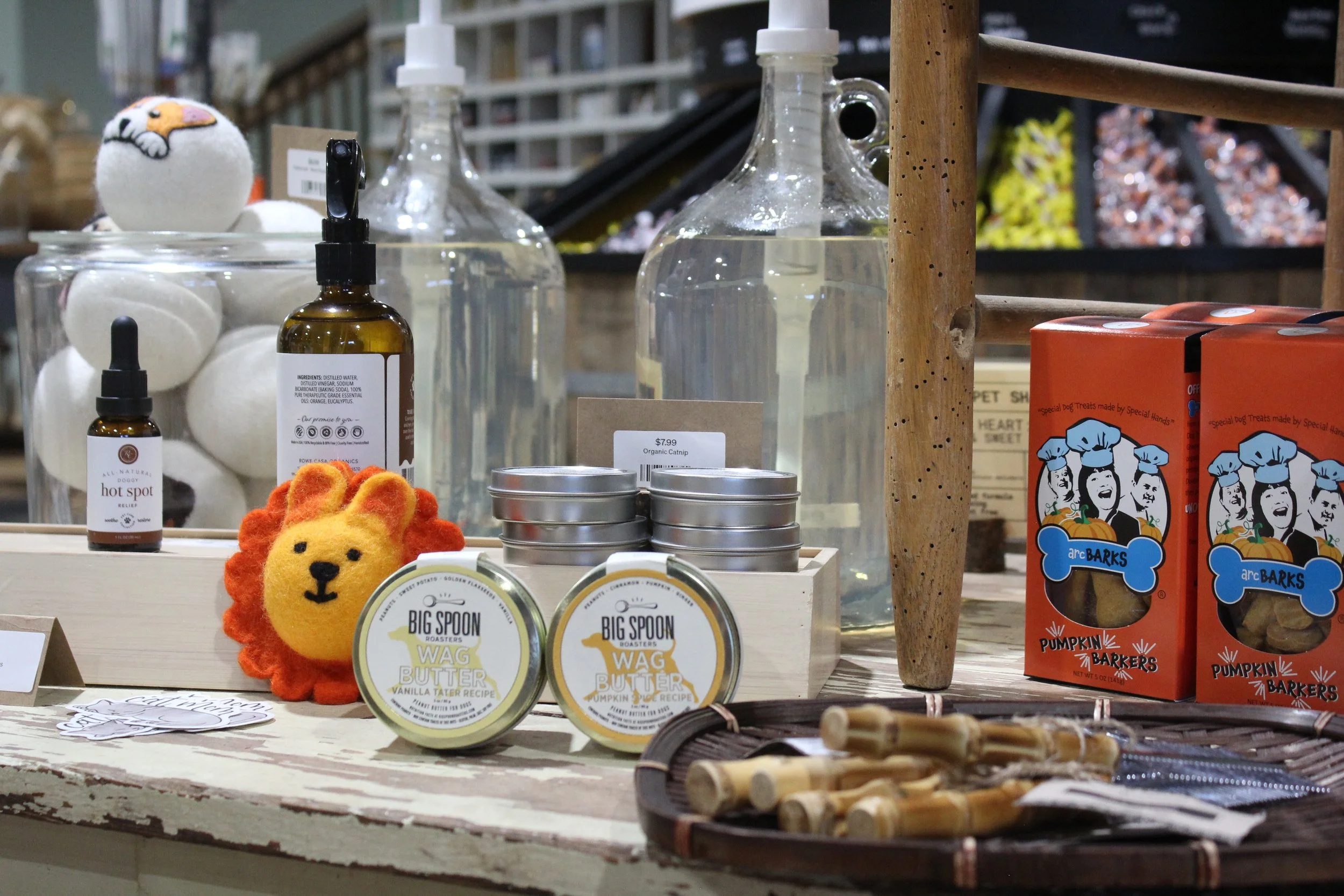 Display of pet products including plush lion toy, bottles of skincare or pet care products, tin containers labeled Wag Butter with pumpkin flavor, and boxes of Pumpkin Barkers dog treats, at Burley Sustainable General Store in West Jefferson, NC.