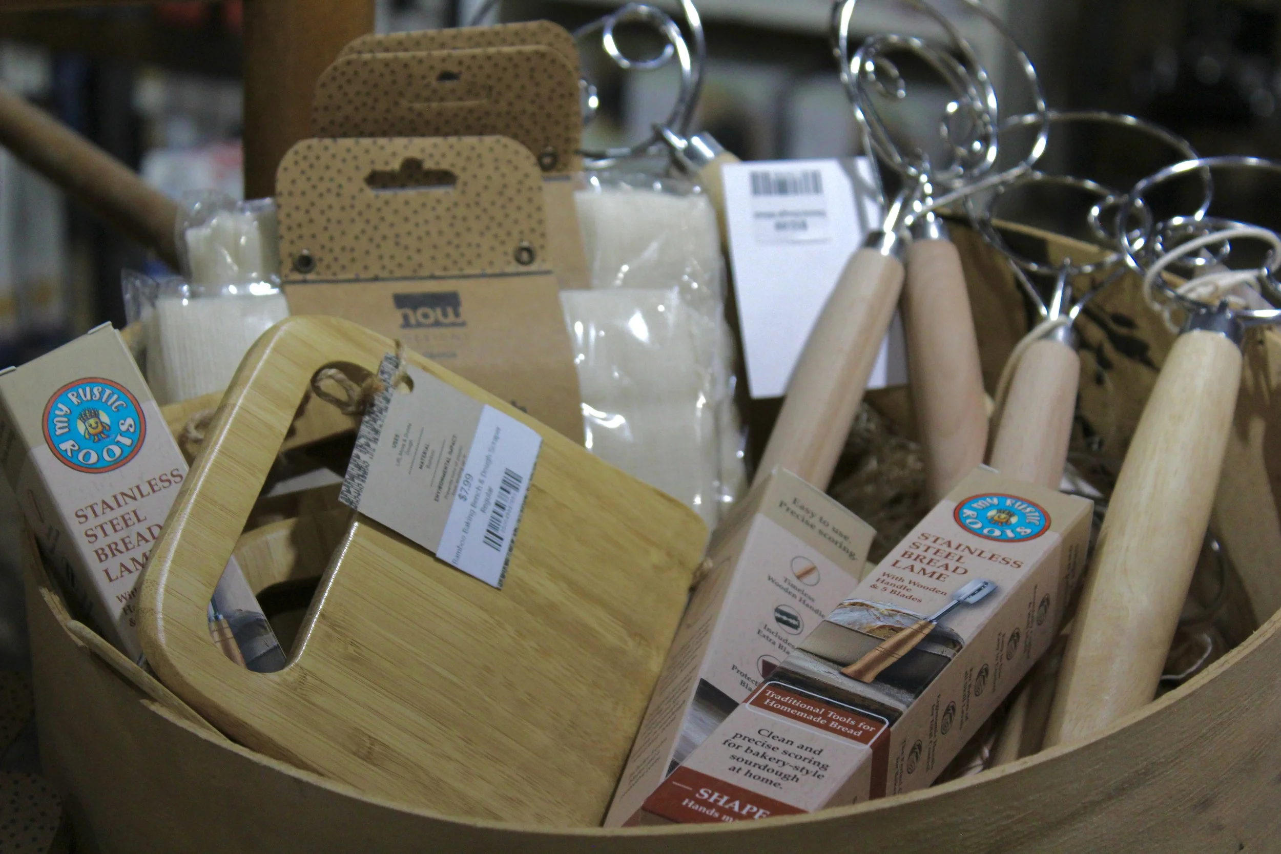 A display of stainless steel bread lames with wooden handles, packaged in clear plastic and cardboard tags, along with other kitchen tools.