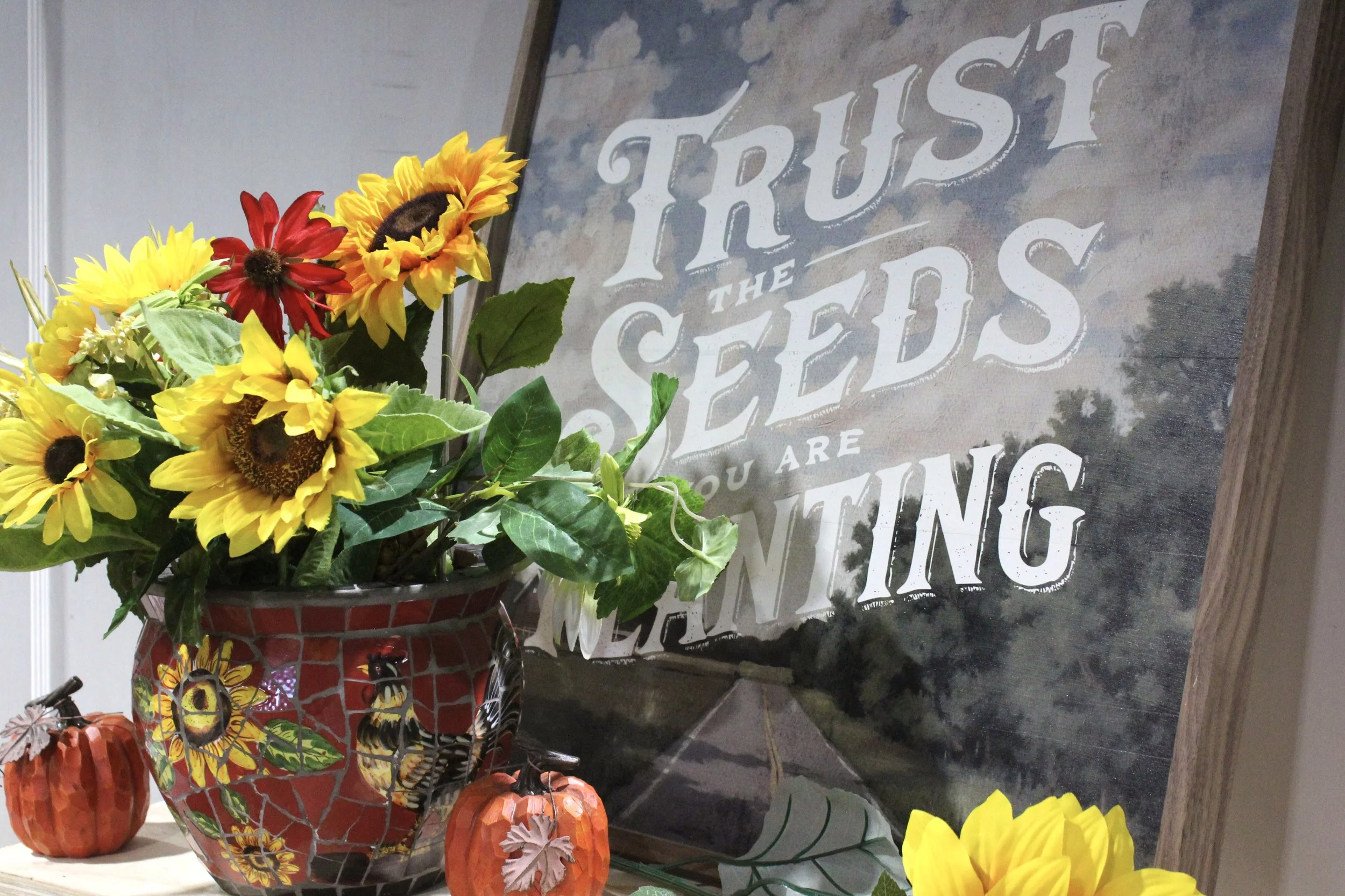 A bouquet of yellow, red, and sunflower flowers in a patterned vase, placing on a table with small pumpkin decorations, beside a decorative sign with white text on a weathered background.