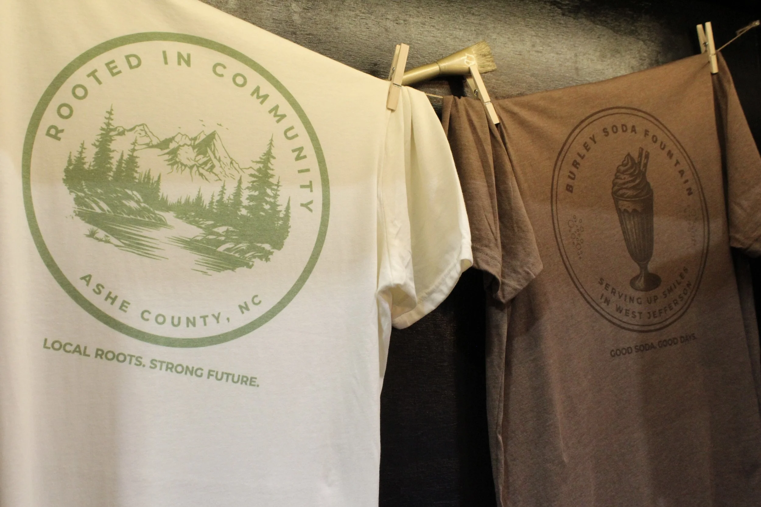 100% cotton t shirts with 'Rooted in Community Ash County, NC, Local Roots. Strong Future.' and another with a design of a soda float and the text 'Buyery Soda Fountain Serving Up Good Days in West Jefferson.'.