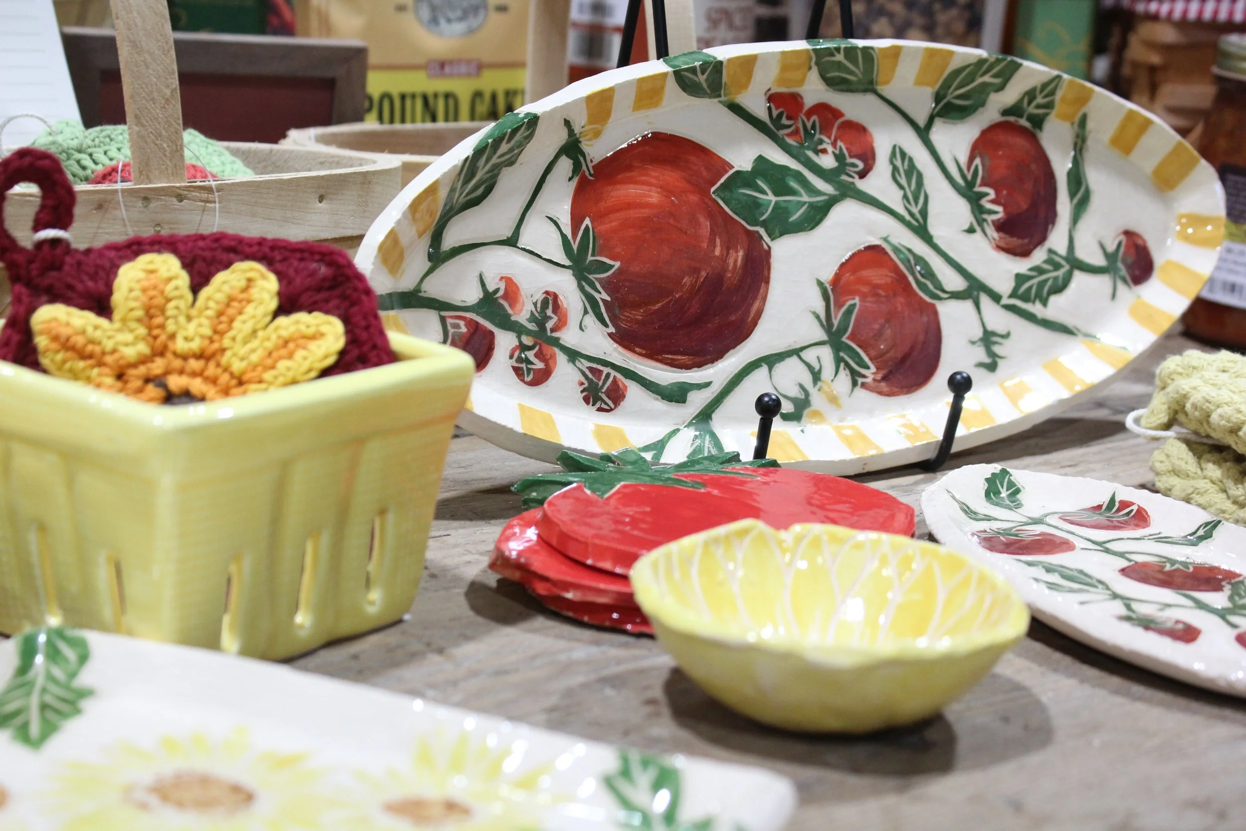 A colorful display of ceramic pottery and dishware, featuring a large white tray with painted red tomatoes and green vines, small bowls patterned with yellow lemons, and a yellow mug with embroidered orange and yellow flowers.