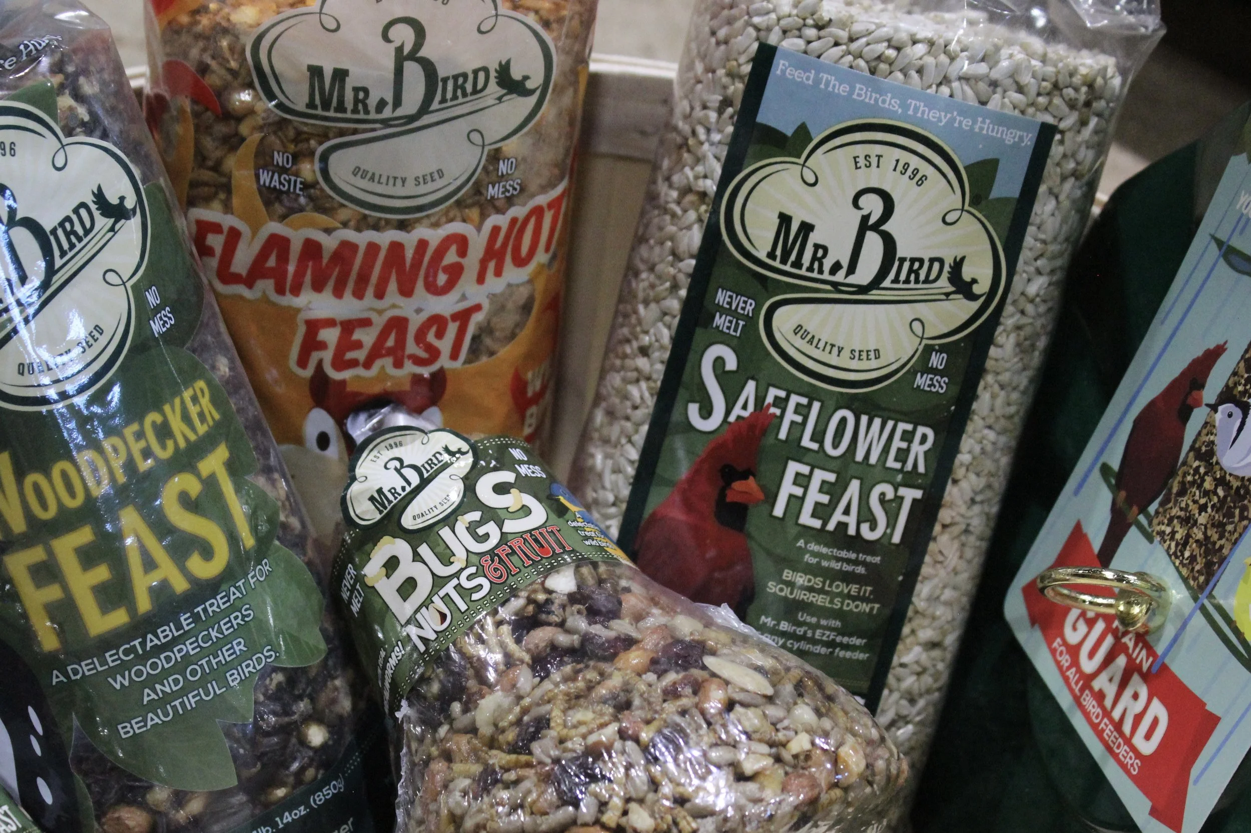 Various bird seed packages on display, including brands like Mr. Bird with labels indicating they contain quality seed and are no waste or mess. The packages include labels like 'Flaming Hot Feast,' 'Saflower Feast,' and a mix of nuts and fruits for 