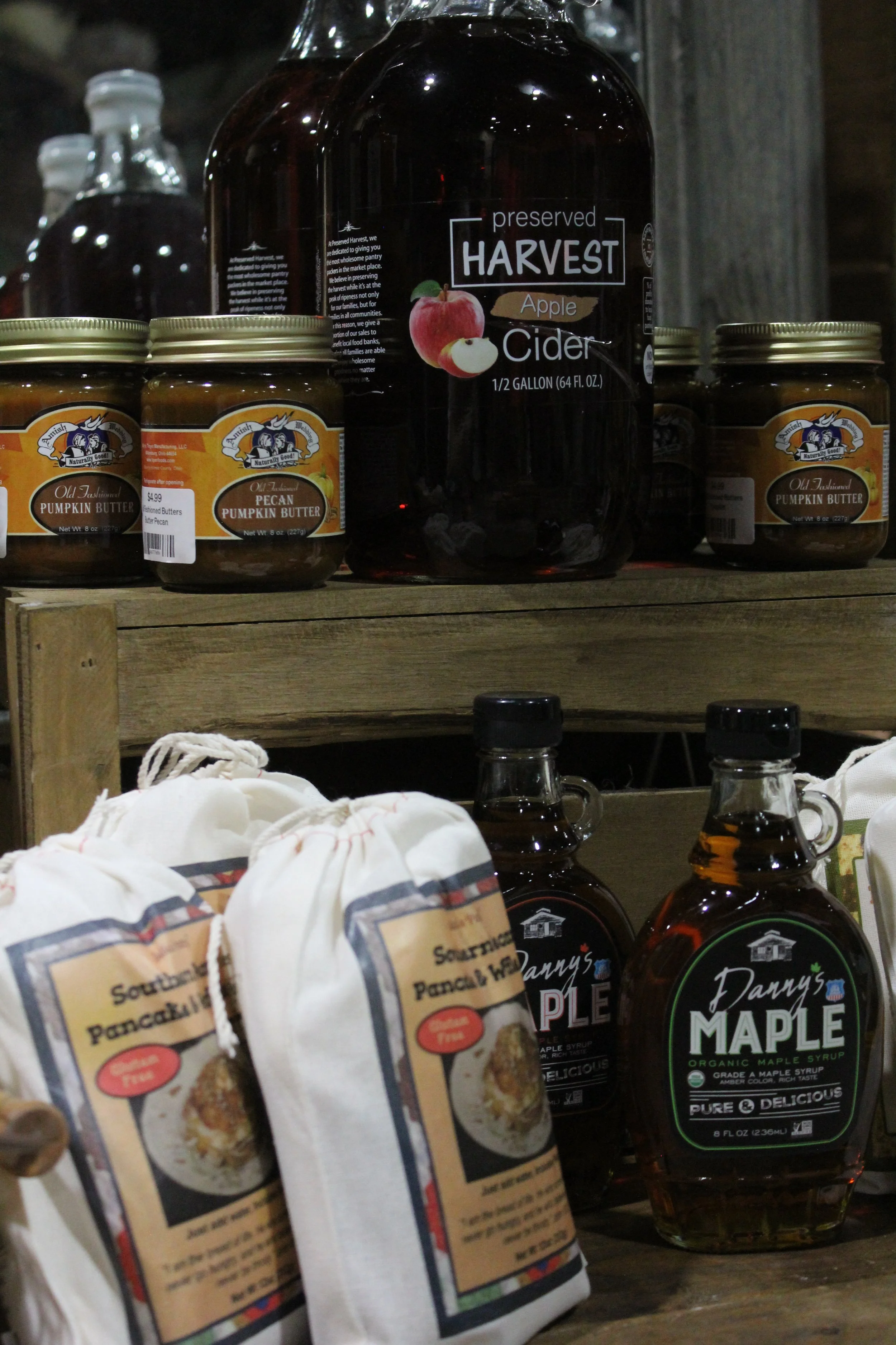 A shelf displaying jars of pumpkin butter, bottles of maple syrup, and a large bottle of apple cider.