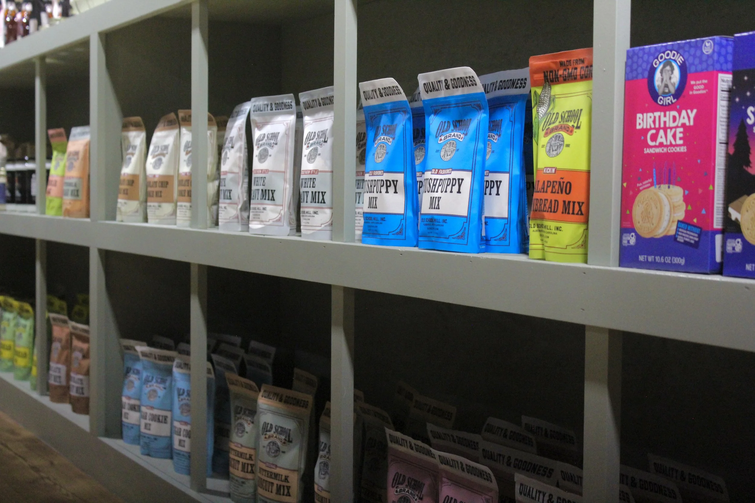 Shelf with various flavored cookie and snack packages, including Old School Brand Pups Puppy Mix, birthday cake cookies, and jalapeno cornbread mix.