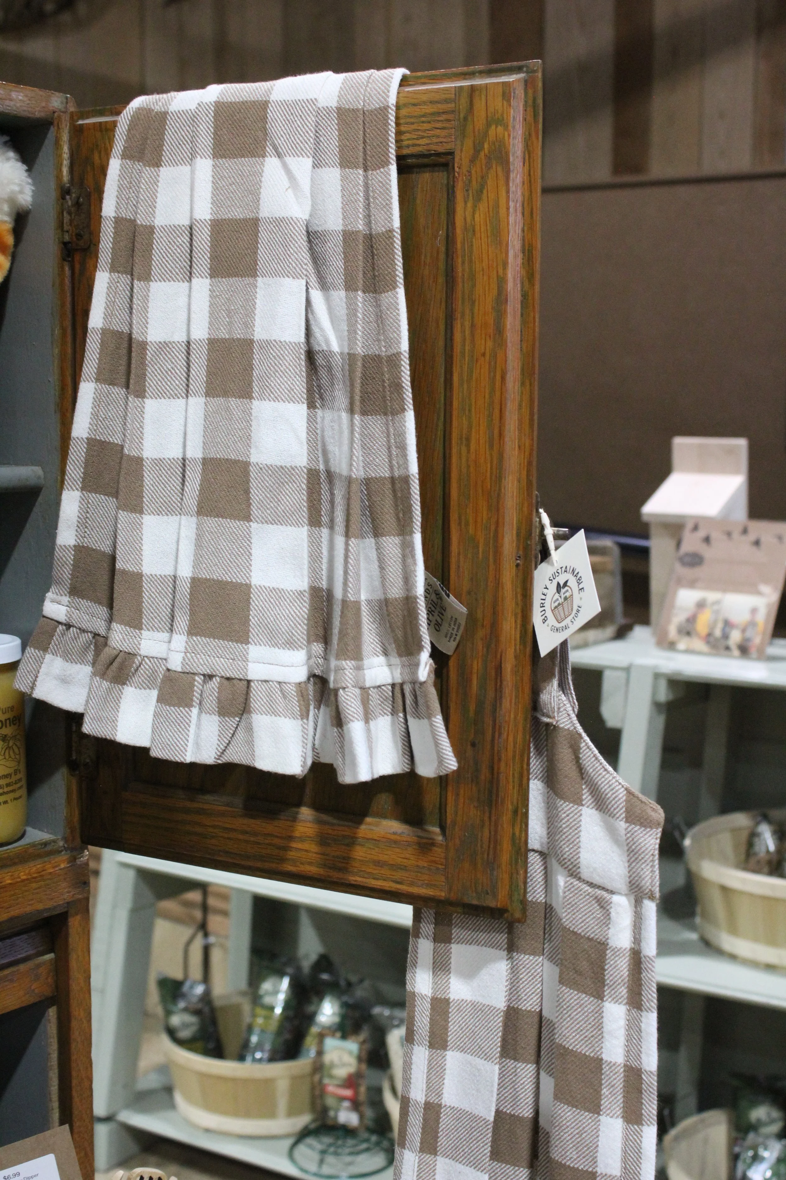 A brown and white checkered kitchen towel draped over a wooden display stand in a store. In the background, there are shelves with various household items including baskets, bottles, and tools.