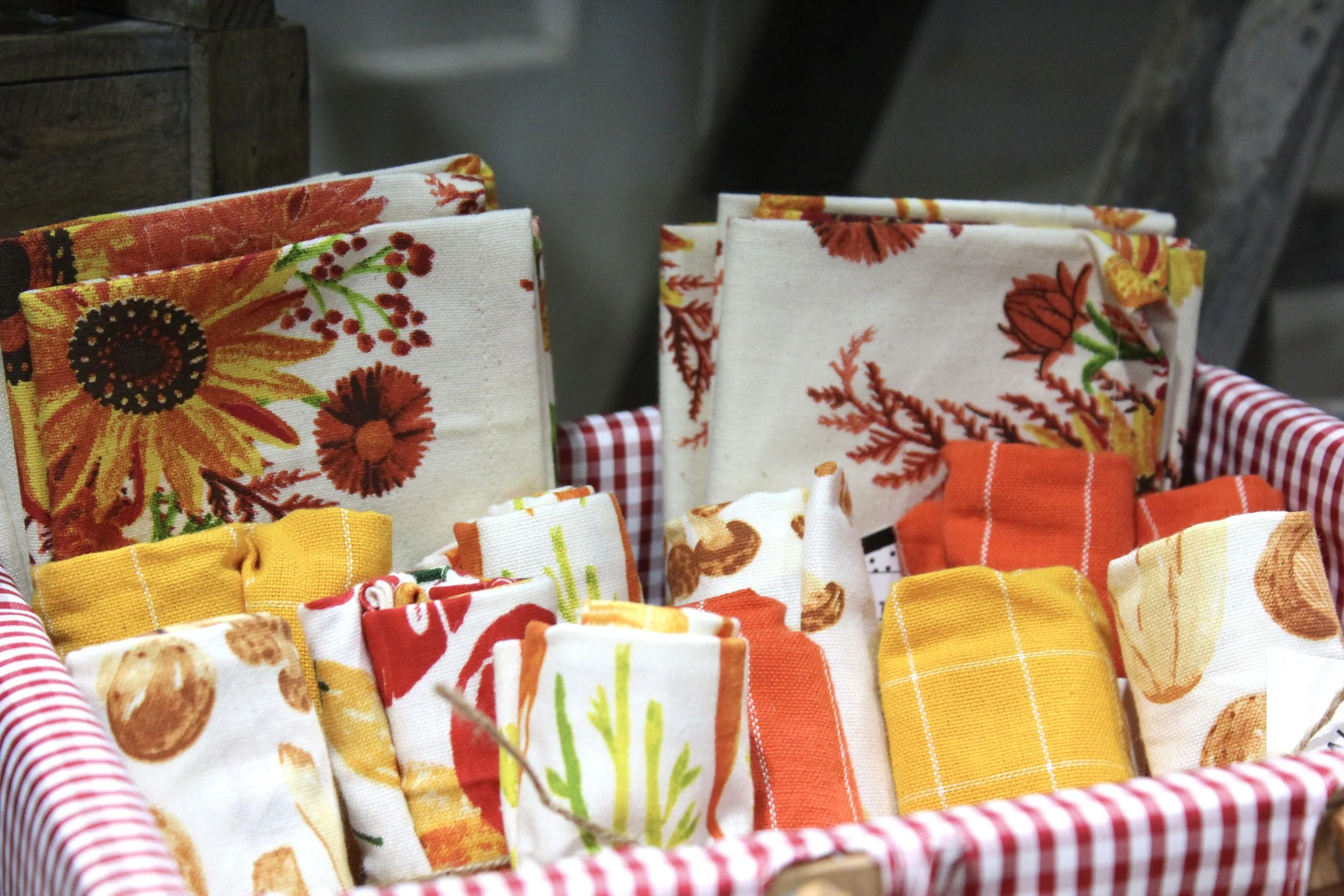 A display of folded fabric napkins with vibrant, floral, and fall-inspired patterns in a basket with a red and white striped fabric lining.