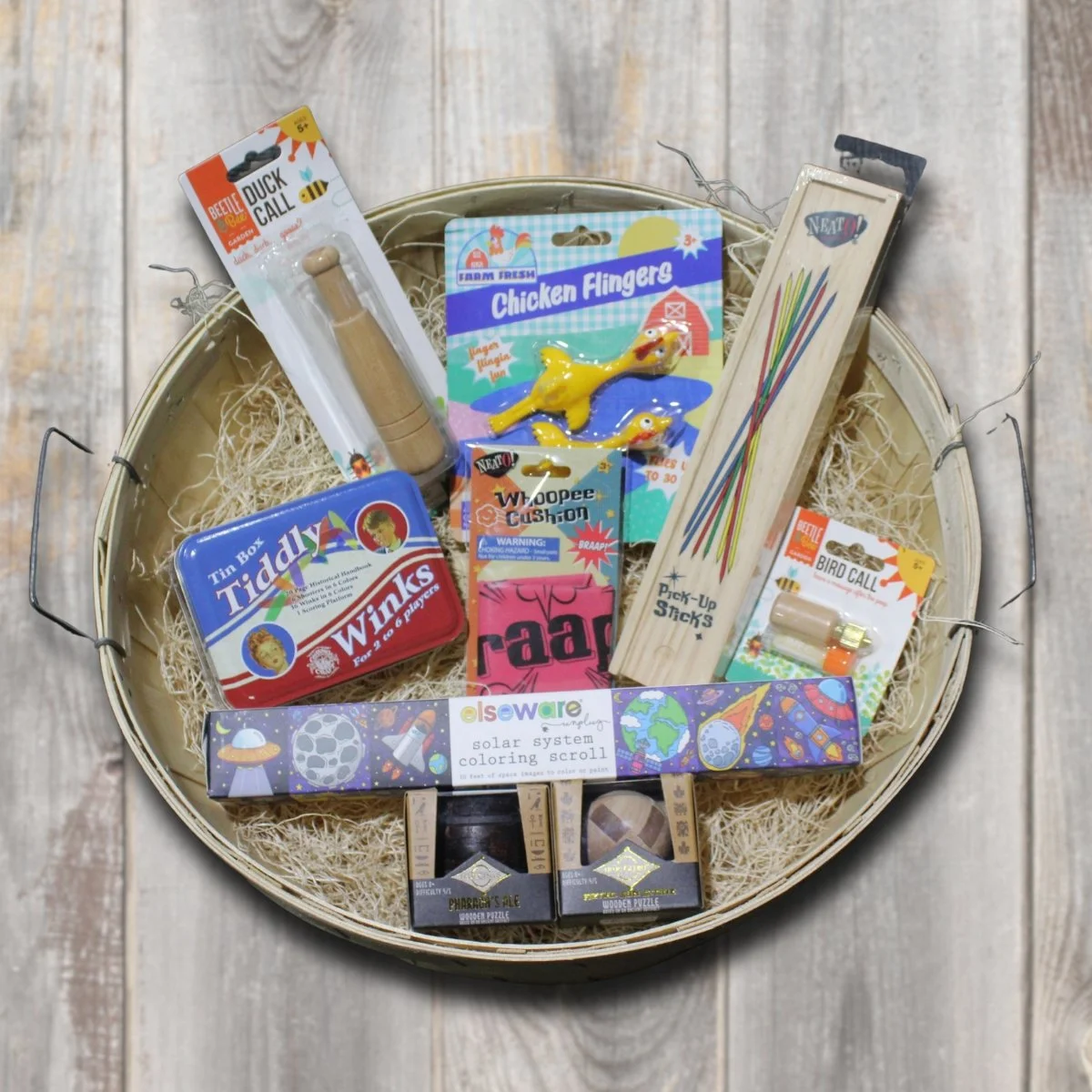 A round basket filled with children's toys and games, including a wooden duck call, chicken flingers rubber ducks, a pick-up sticks game, a bird call, a solar system coloring scroll, a toy egg-shaped figure, and a mini puzzle.