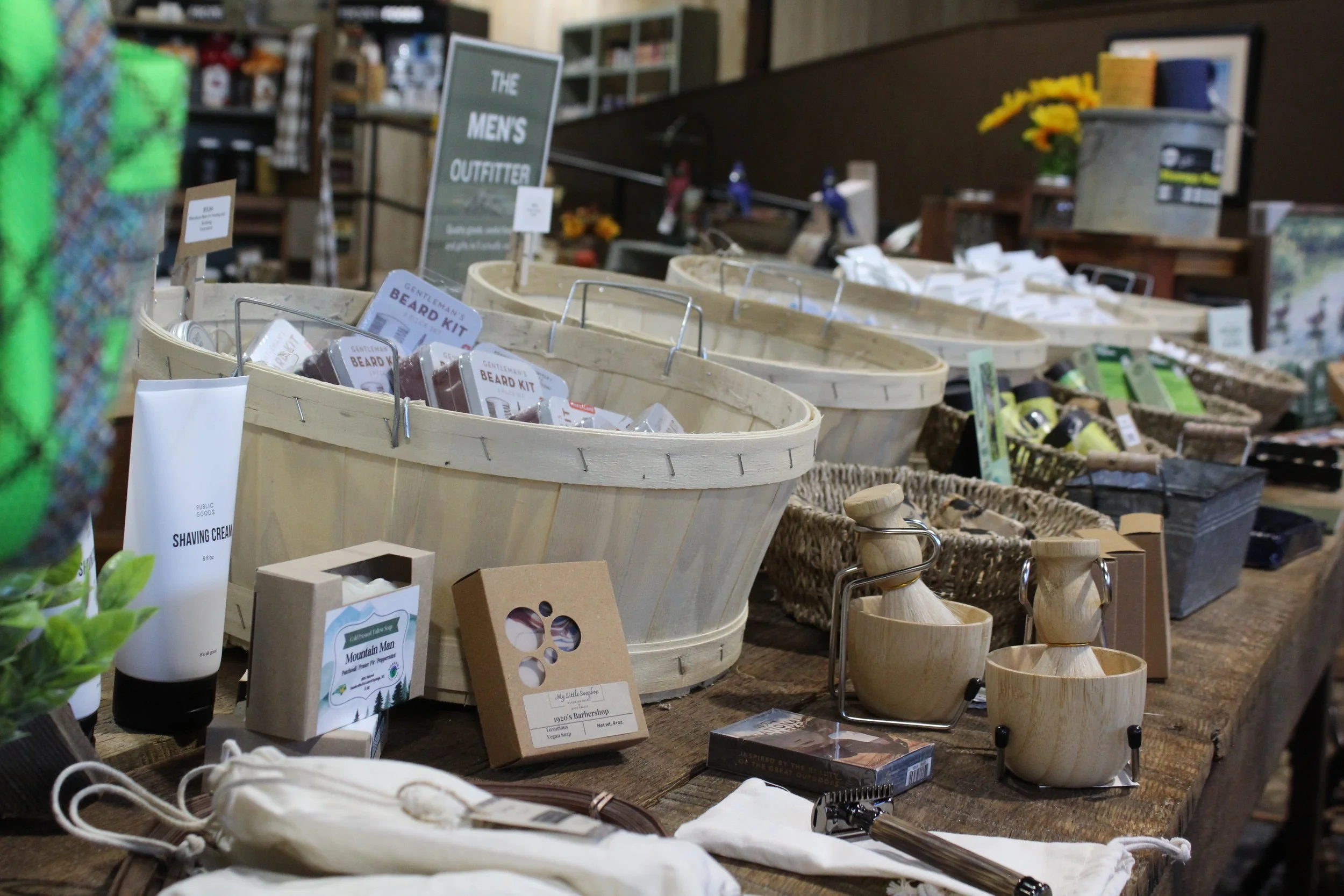 Various outdoor and camping-themed products at Burley Sustainable General Store in West Jefferson, NC, including wooden utensils, soap, skincare, and beard kit packages, with a sign indicating men's outdoor apparel.