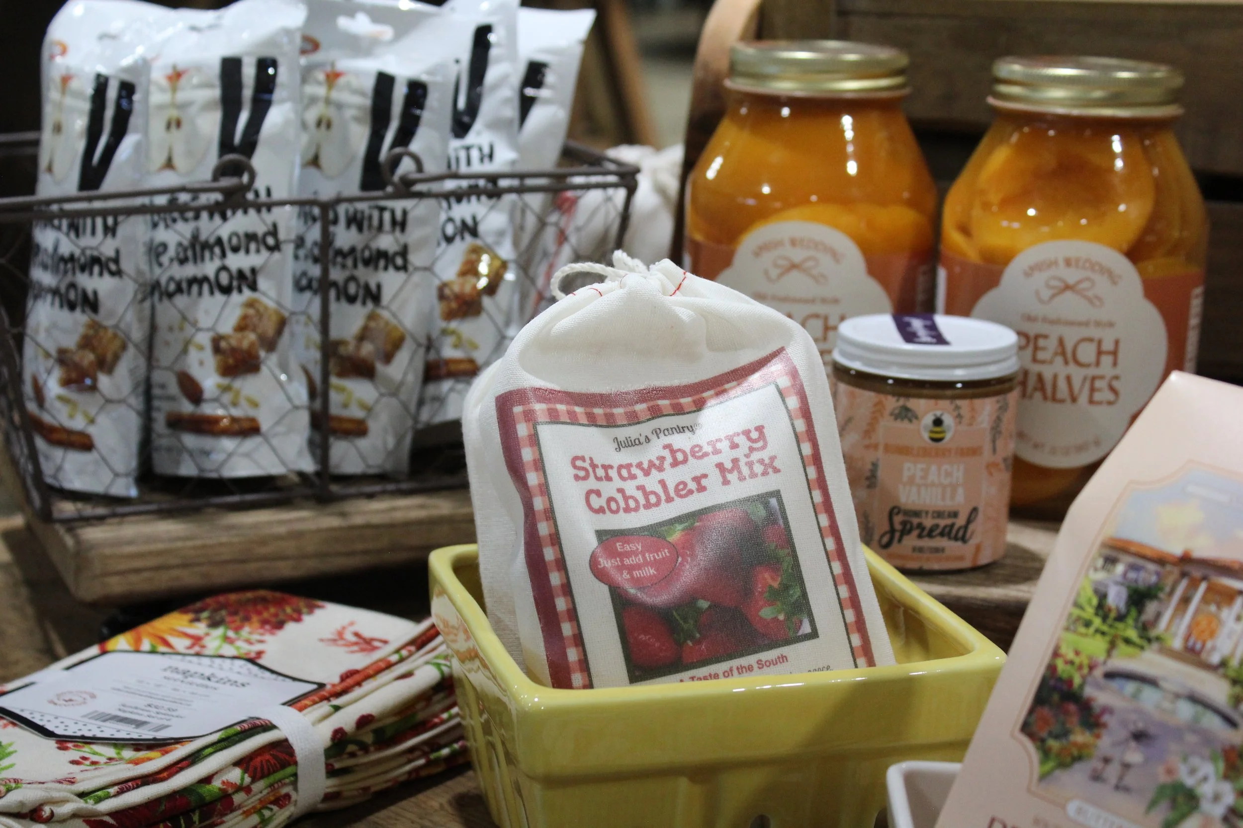 Assorted jars of peach preserves, a bag of strawberry cobbler mix, and a basket of folded holiday-themed napkins on display at Burley Sustainable General Store in West Jefferson, NC.