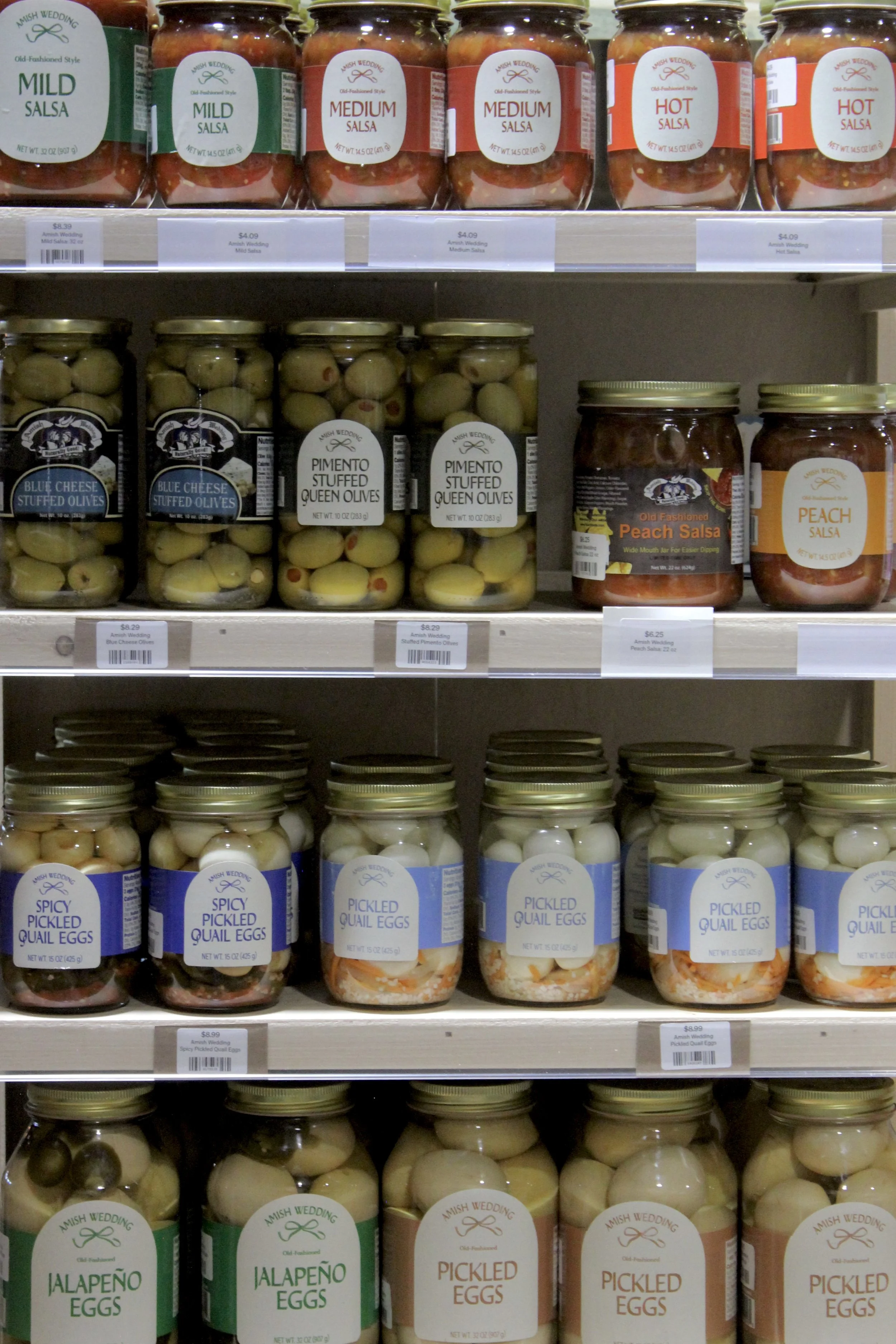 Shelf display of various jars of pickled foods including salsa in different heat levels, stuffed olives, peach salsa, and pickled eggs with labels indicating flavors like mild, medium, hot, jalapeño, and spicy pickled quail eggs.