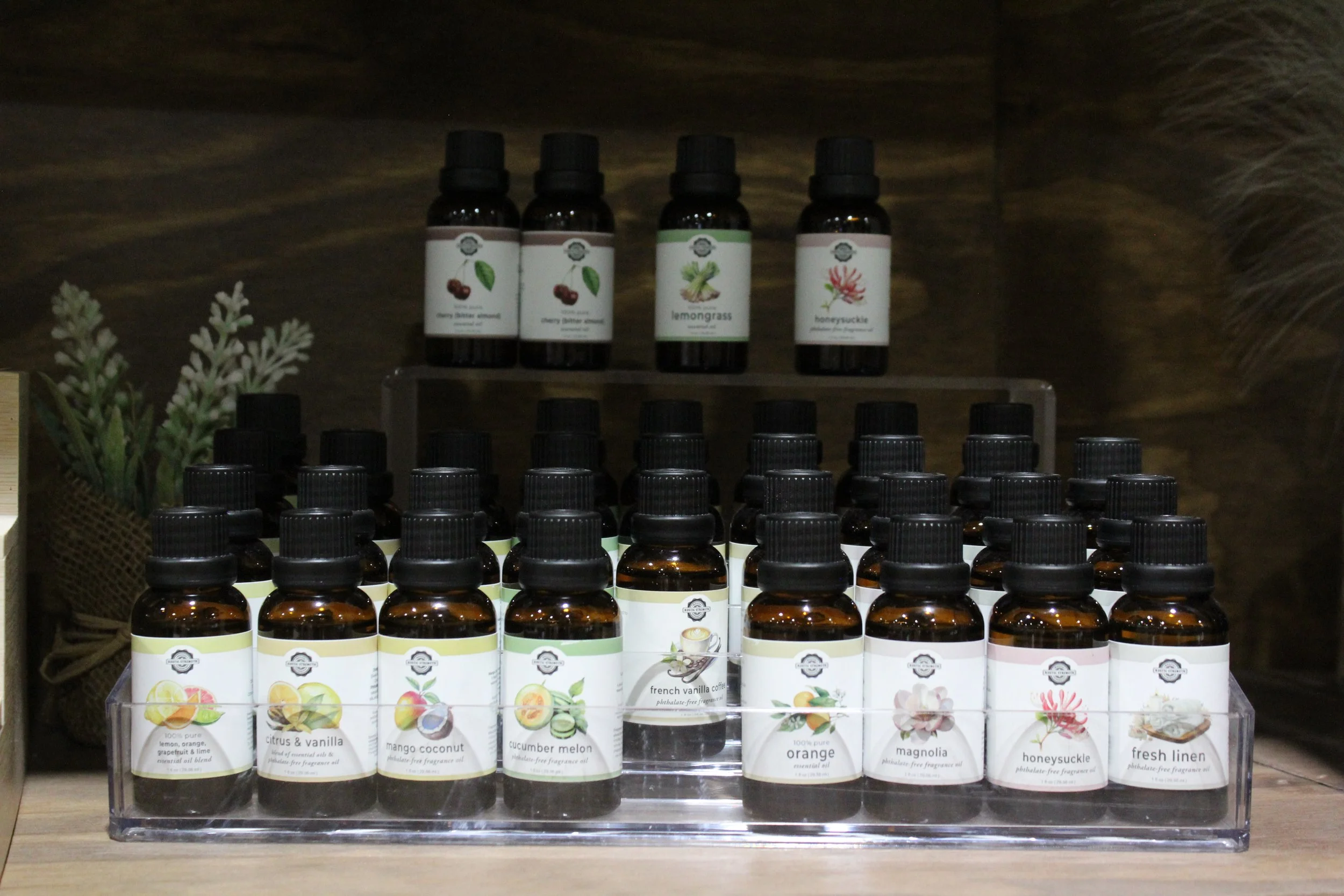 Multiple small bottles of essential oils displayed on a clear acrylic stand, with labels featuring different scents such as citrus & vanilla, mango coconut, cucumber melon, and French vanilla coffee Burley Sustainable General Store.