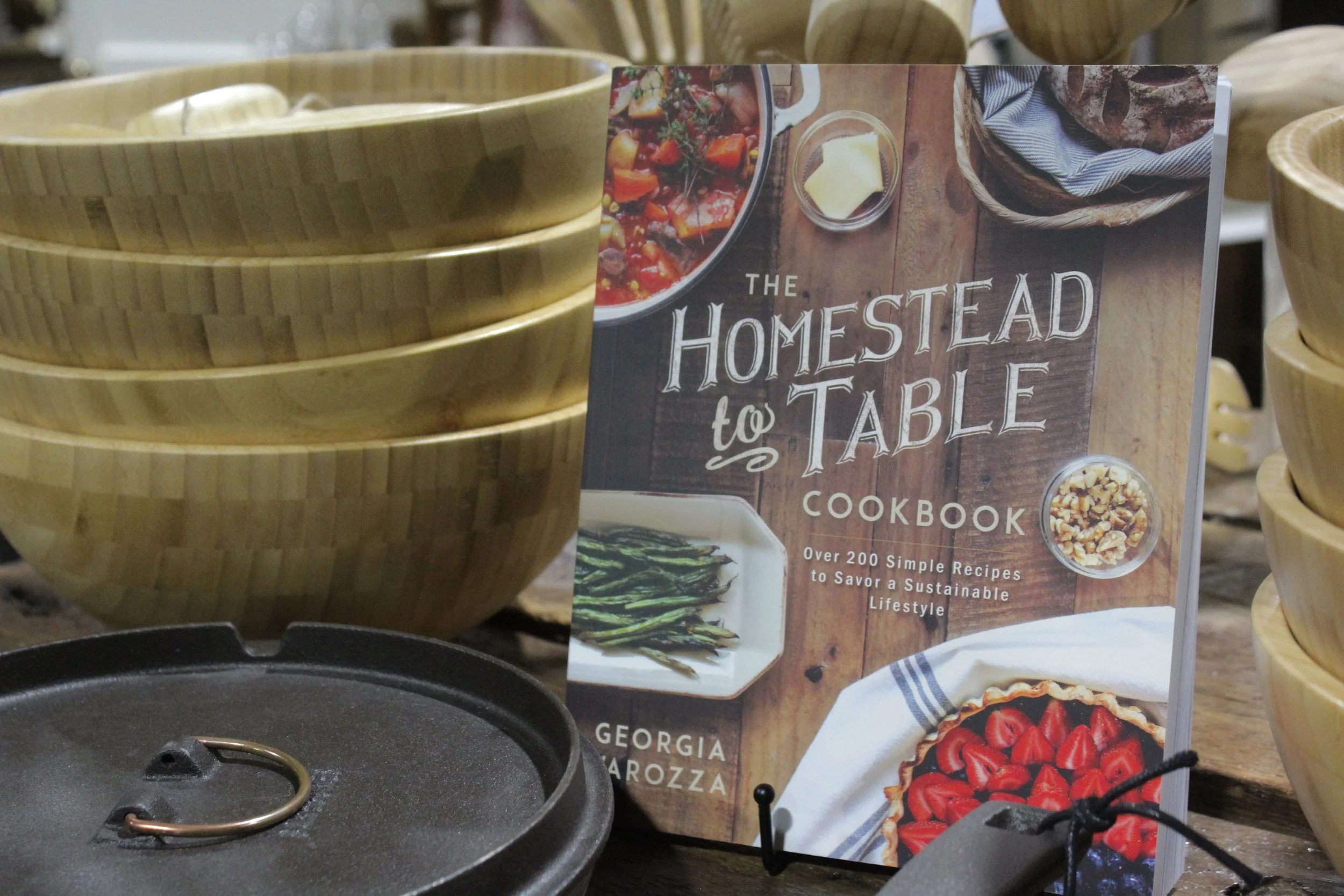 A cookbook titled "The Homestead to Table Cookbook" displayed upright among wooden bowls and a cast iron skillet. 