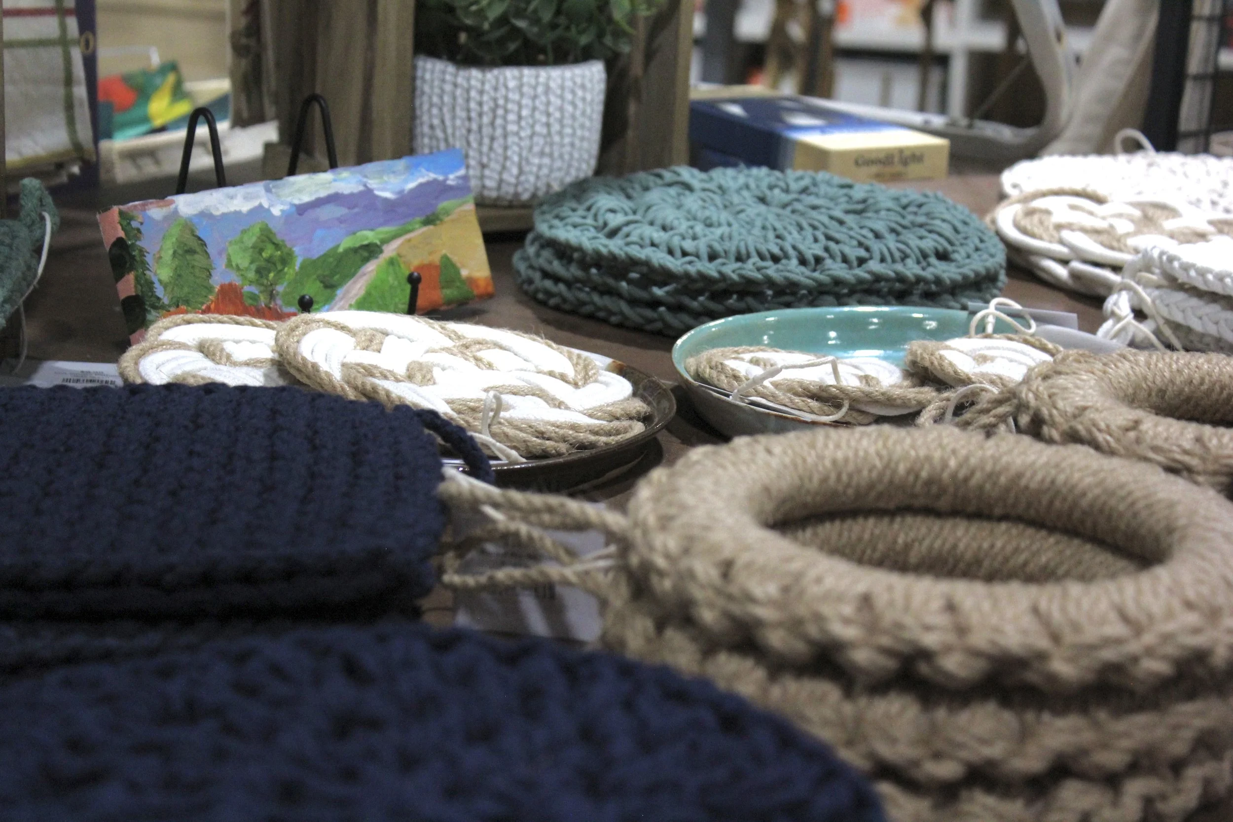 Display of handmade knitted and woven items, including ropes and hats, on a table with a landscape painting and potted plant in the background.