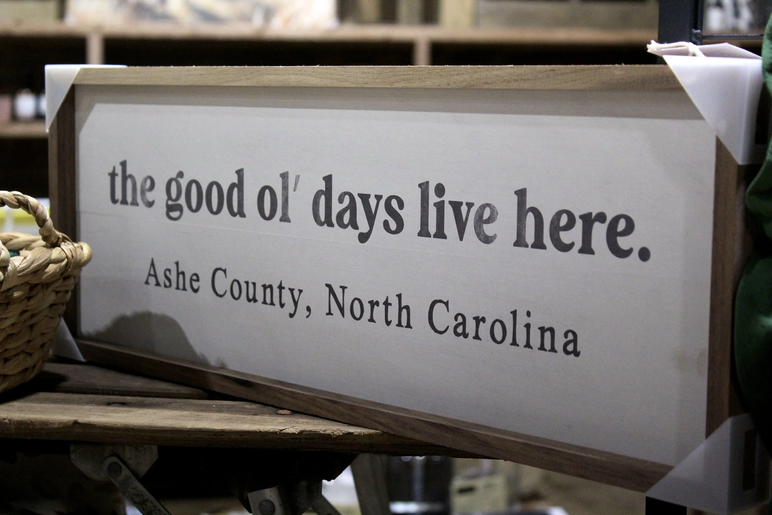 A wooden sign with gray text that reads: "the good ol' days live here." The sign is on a cart at Burley Sustainable General Store in West Jefferson, NC.