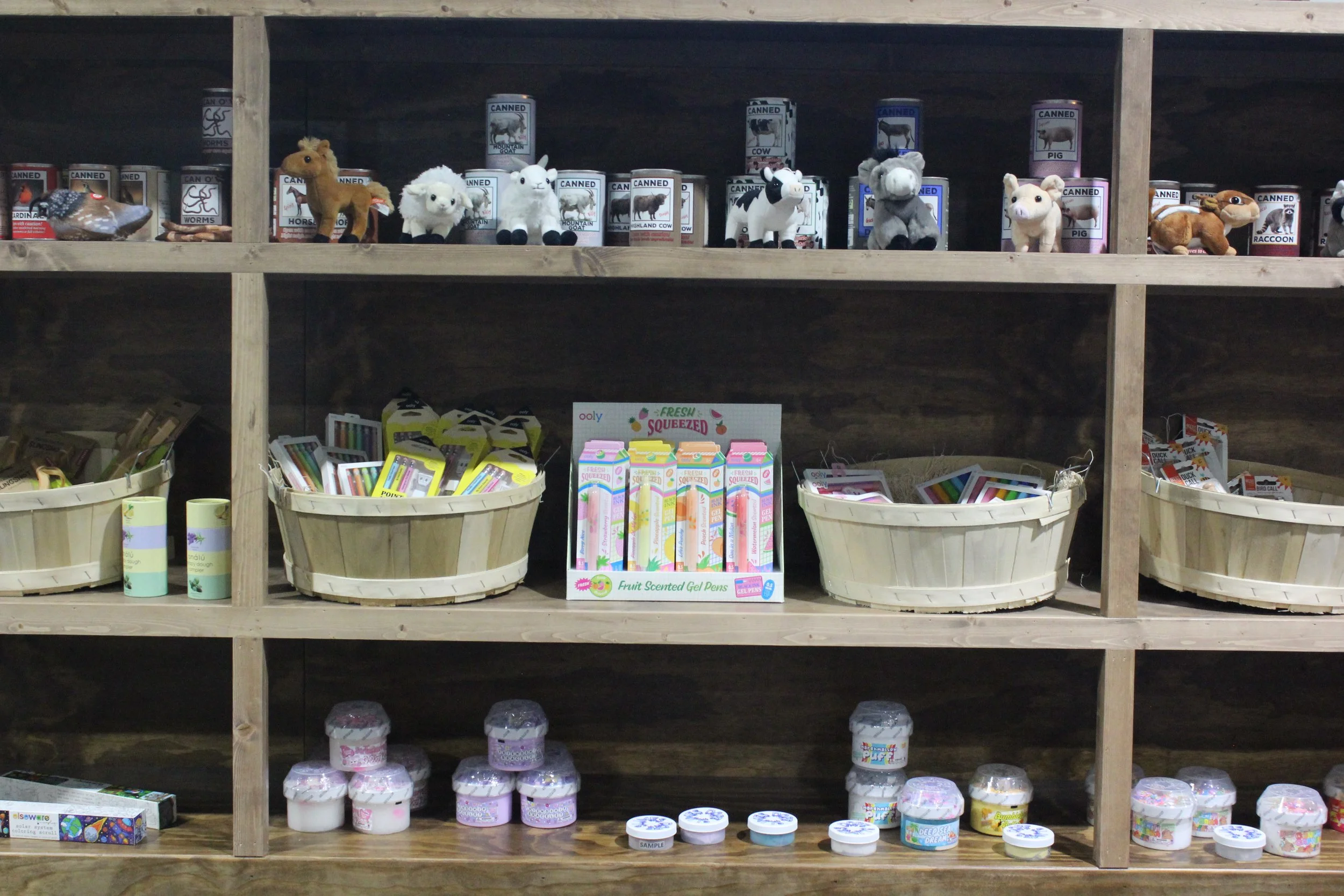 Wooden shelves displaying canned crittes plush animal toys, including a horse, sheep, llama, cow, pig, raccoon, and squirrel,  baskets of colorful gel pens and outdoor toys for kids.